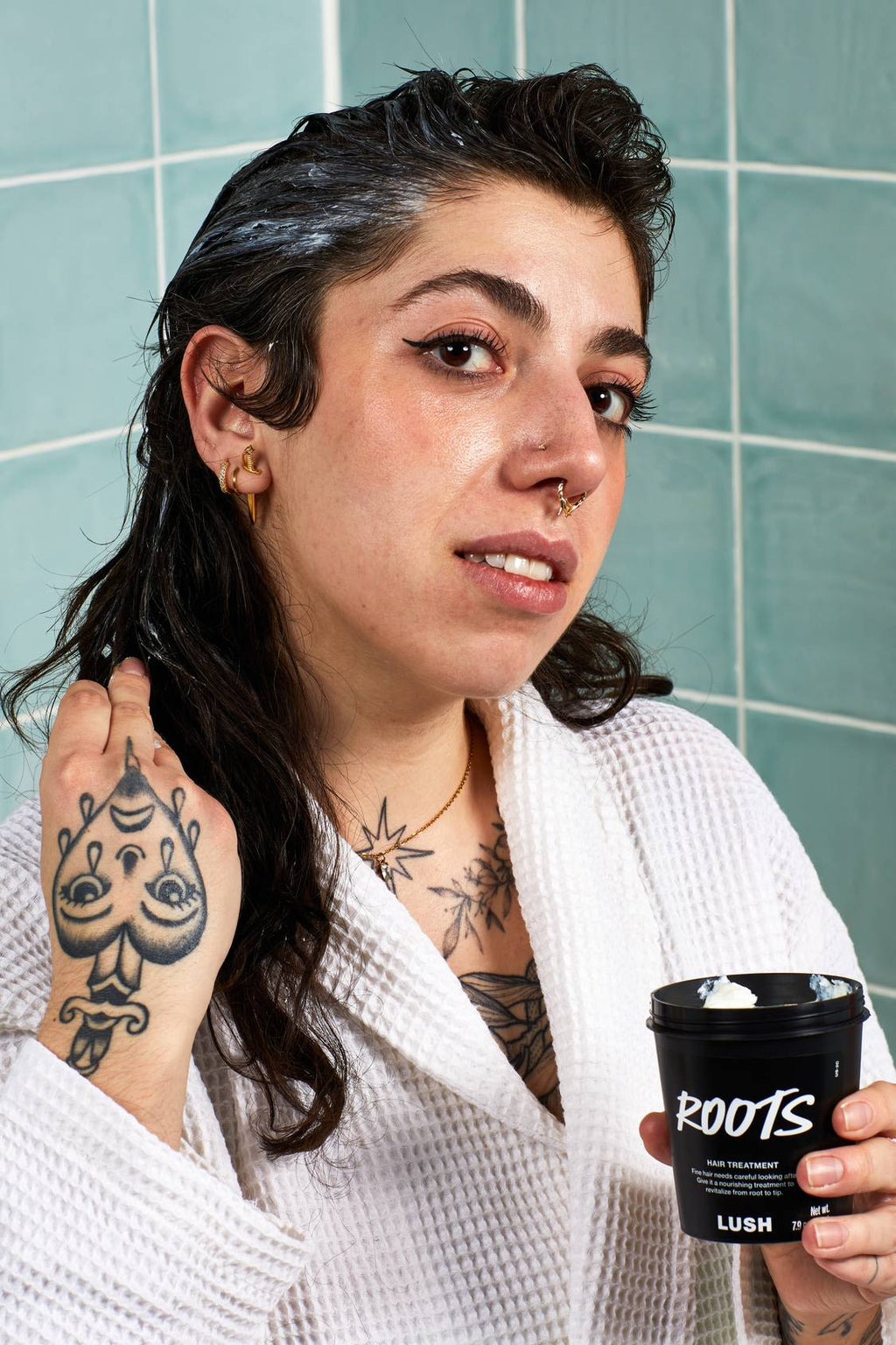 A person wearing a white bathrobe holds a black pot of Lush Roots hair treatment while massaging the white cream into their dark, wet hair. They have tattoos on their chest and hand, set against a background of blue-green tiles.