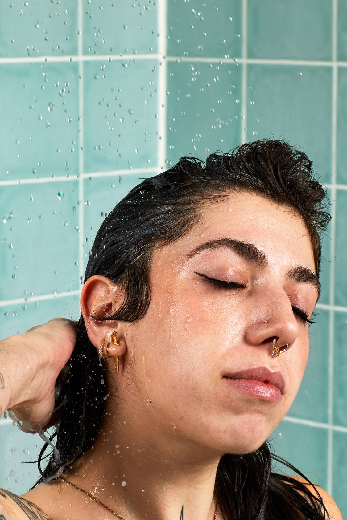 A close-up lifestyle shot of a person with dark hair tilting their head back with eyes closed under a spray of running shower water. They are rinsing their hair against a background of light blue-green tiles.