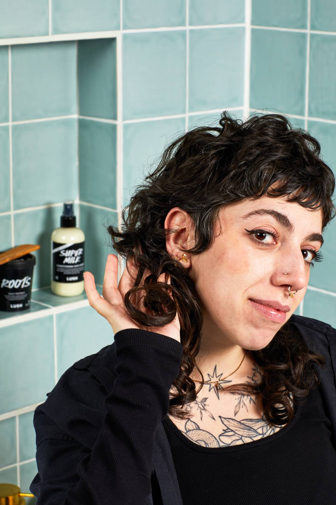 A smiling person scrunges their bouncy, dark curly hair. In the background shelf of the blue-green tiled bathroom, a bottle of Lush Super Milk conditioning spray and a black pot of Roots hair treatment are visible.