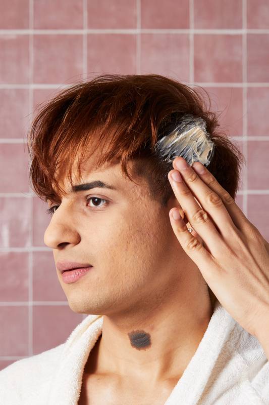 A model with copper-coloured hair and a white robe applies the creamy, LUSH "H'suan Wen Hua" hair treatment to their hair with their fingers. They are shown in profile against a background of pink square tiles.