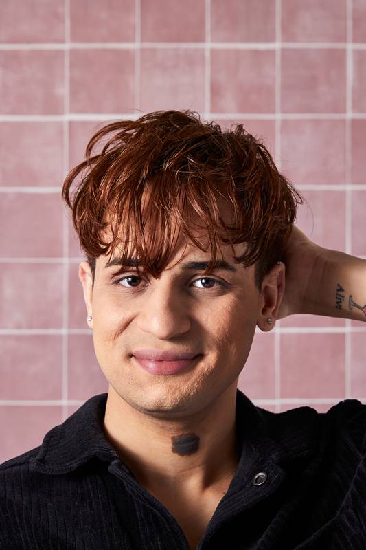 A model with shining, copper-toned hair and a black corduroy shirt smiles at the camera, showing the effects of using the "H'suan Wen Hua" hair treatment. They are positioned against a background of soft pink square tiles.
