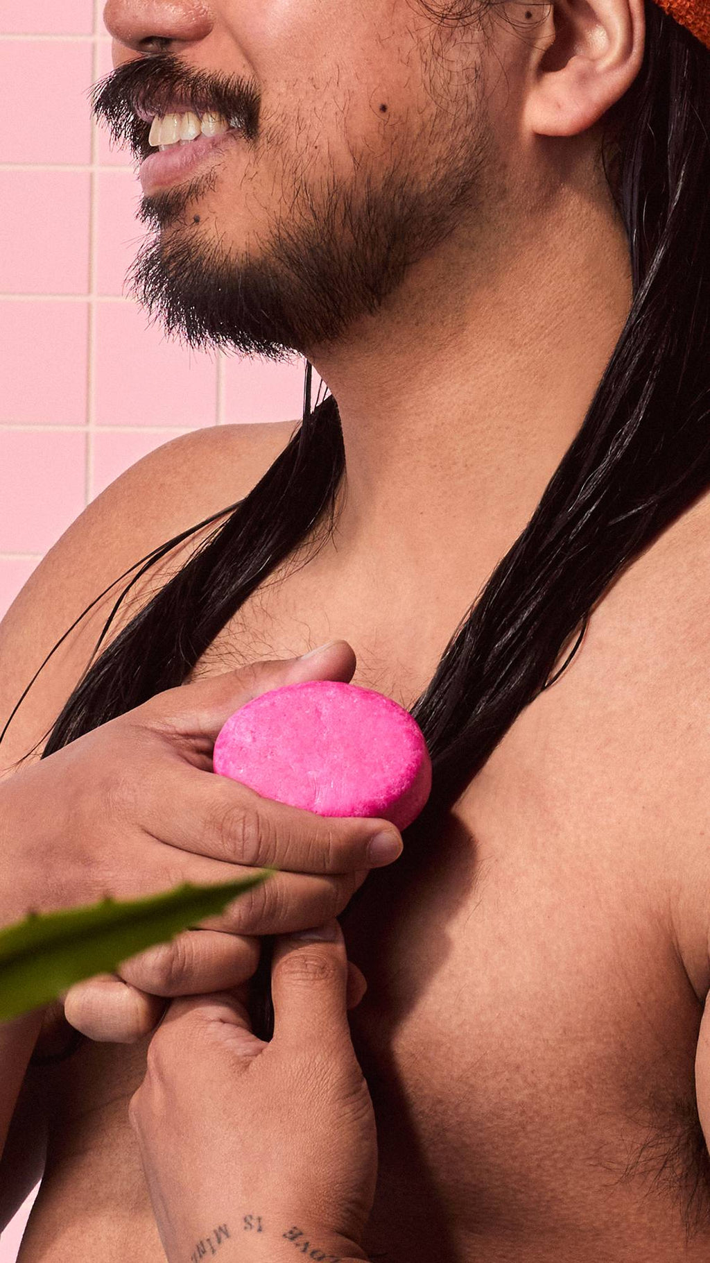 Close-up of a smiling person's lower face and torso as they rub a pink Lush "American Cream" solid conditioner bar down a section of wet dark hair in a pink-tiled shower.