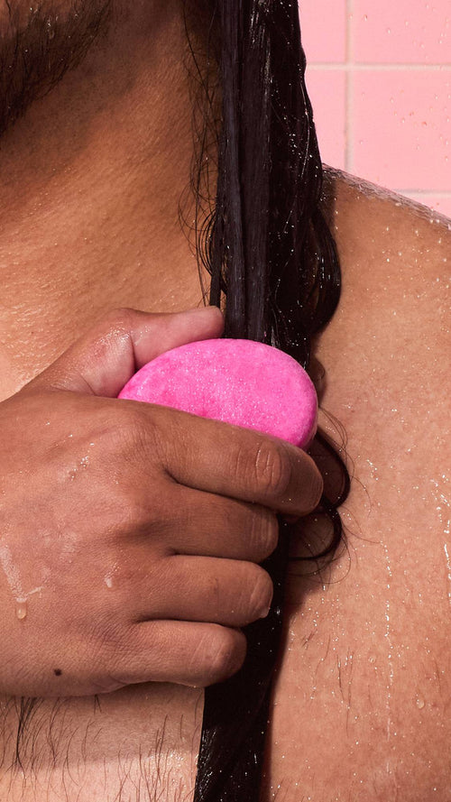 Close-up image of a person smoothing a bright pink solid Lush "American Cream" conditioner bar down a strand of wet, dark hair in the shower against light pink tiles.