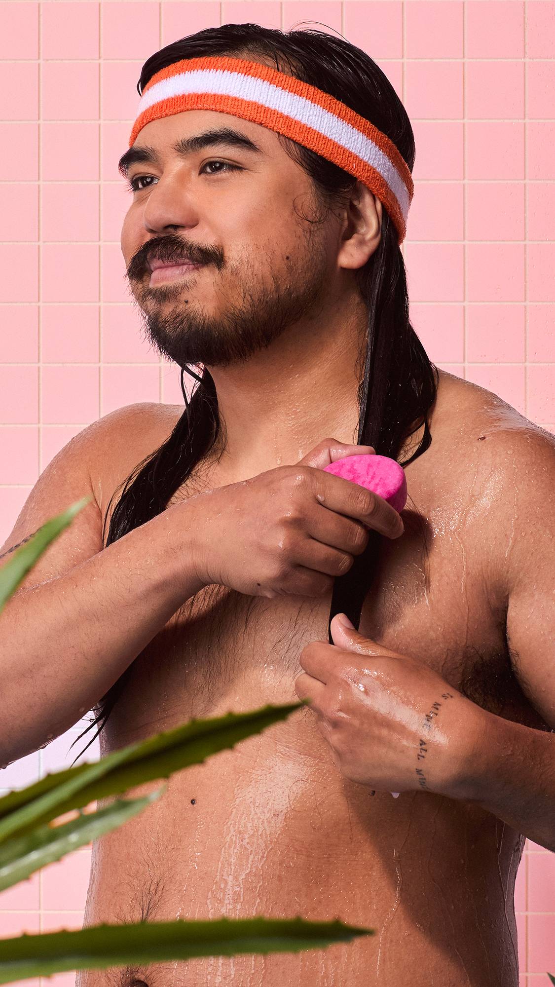 A person with long dark hair and an orange headband runs a pink Lush "American Cream" solid conditioner bar down their hair in a shower with pink tiled walls.