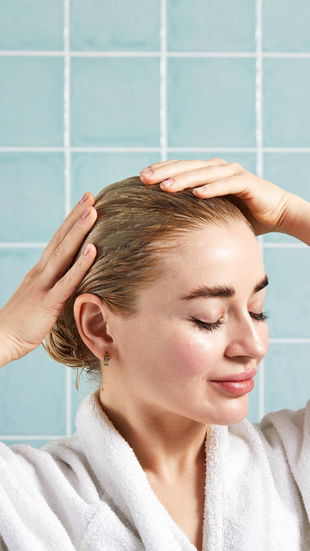 A side-profile view of a smiling model with their eyes closed, massaging the Lush "Marilyn" hair treatment through their damp blonde hair with both hands. They wear a white robe against a background of glossy, light blue square tiles.
