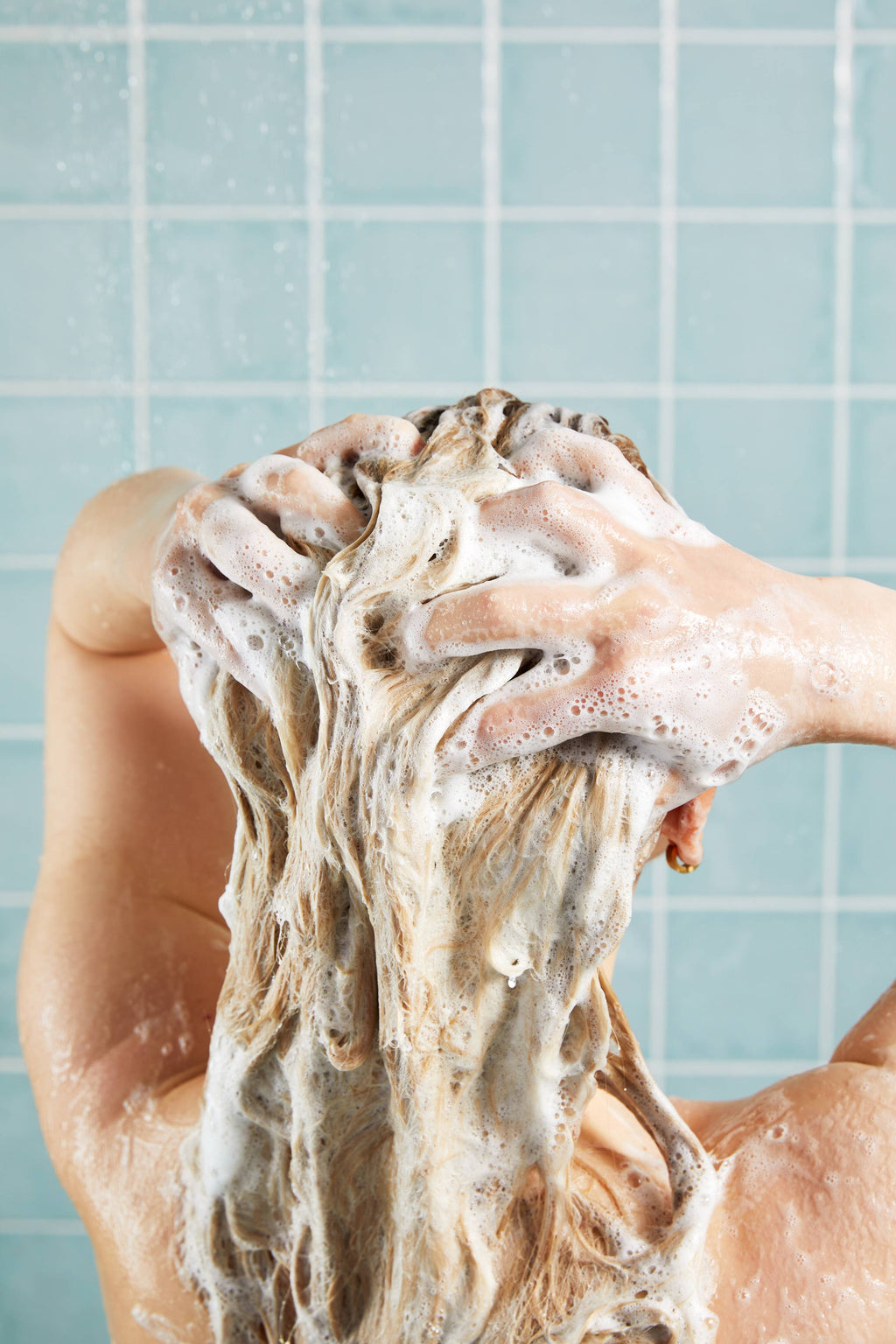 A view from behind of a person with blonde hair, massaging a rich, white shampoo lather through their hair. Drops of water fall around them against a backdrop of pale blue square bathroom tiles.