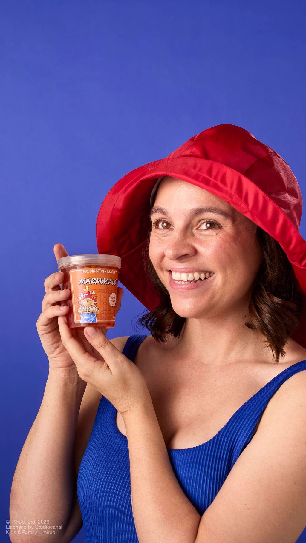 A model with brown hair, wearing a red rain hat and a blue ribbed vest, smiles while holding a pot of 'Marmalade' body wash from the Paddington x Lush range. The pot features an illustration of Paddington Bear. Set against a blue background.