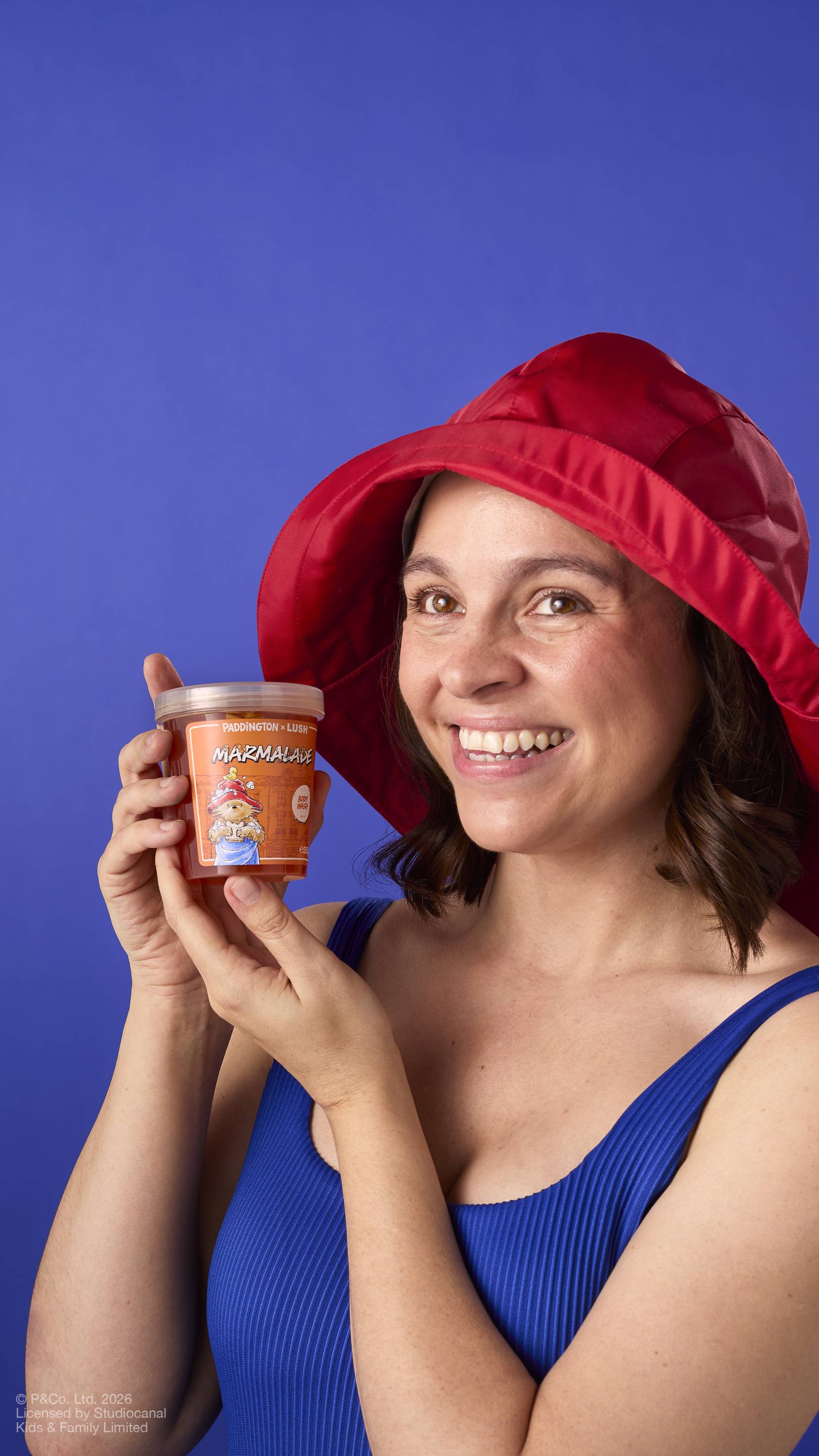 A model with brown hair, wearing a red rain hat and a blue ribbed vest, smiles while holding a pot of 'Marmalade' body wash from the Paddington x Lush range. The pot features an illustration of Paddington Bear. Set against a blue background.