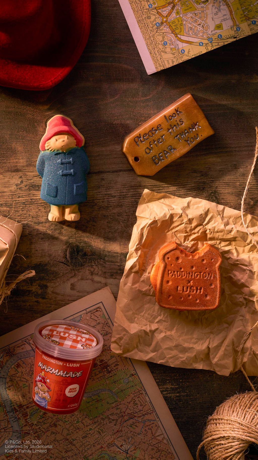 The image shows a collection of Paddington Bear-themed Lush products on a dark wooden surface, including a Paddington bath bomb, a marmalade sandwich-shaped bubble bar, and a 'Marmalade' body wash, around Maps, string, and a red hat.