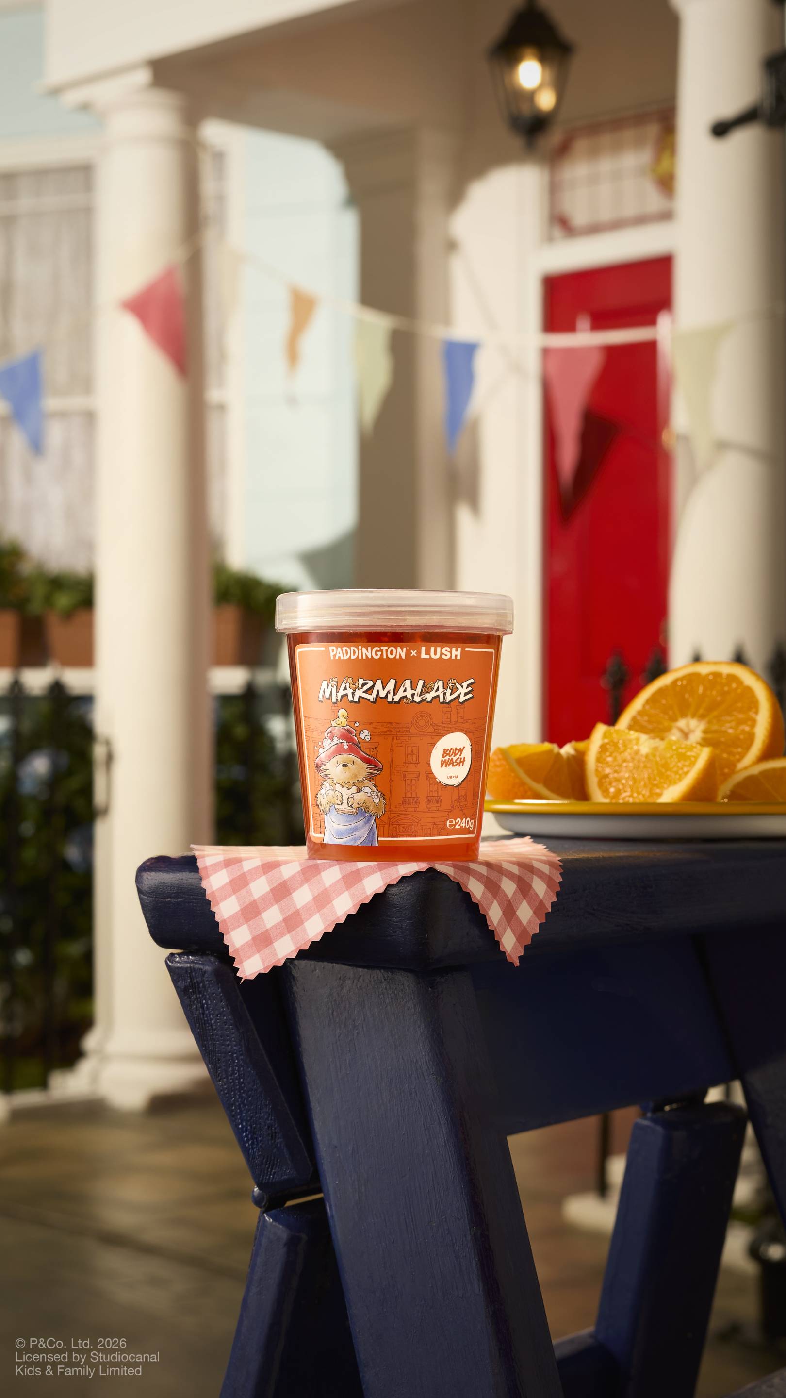 A clear pot of 'Marmalade' body wash from the Paddington x Lush range sits on a blue wooden bench outdoors. The scene includes bright sunshine, a red front door, bunting, and fresh orange slices on a gingham cloth.