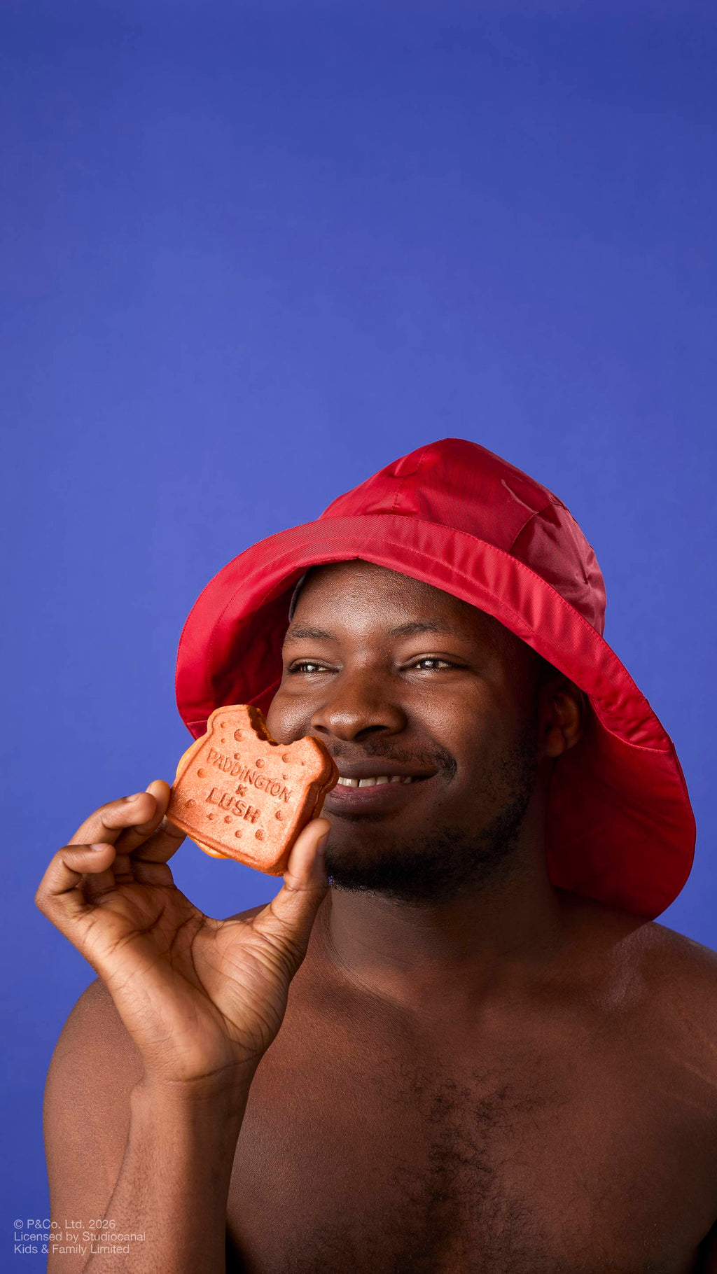 A model in a red rain hat smiles as they hold a "Marmalade Sandwich" bubbleroon bubble bar to their face. The brown, textured bar features a 'bite' and the text 'PADDINGTON x LUSH'. Set against a solid blue background.