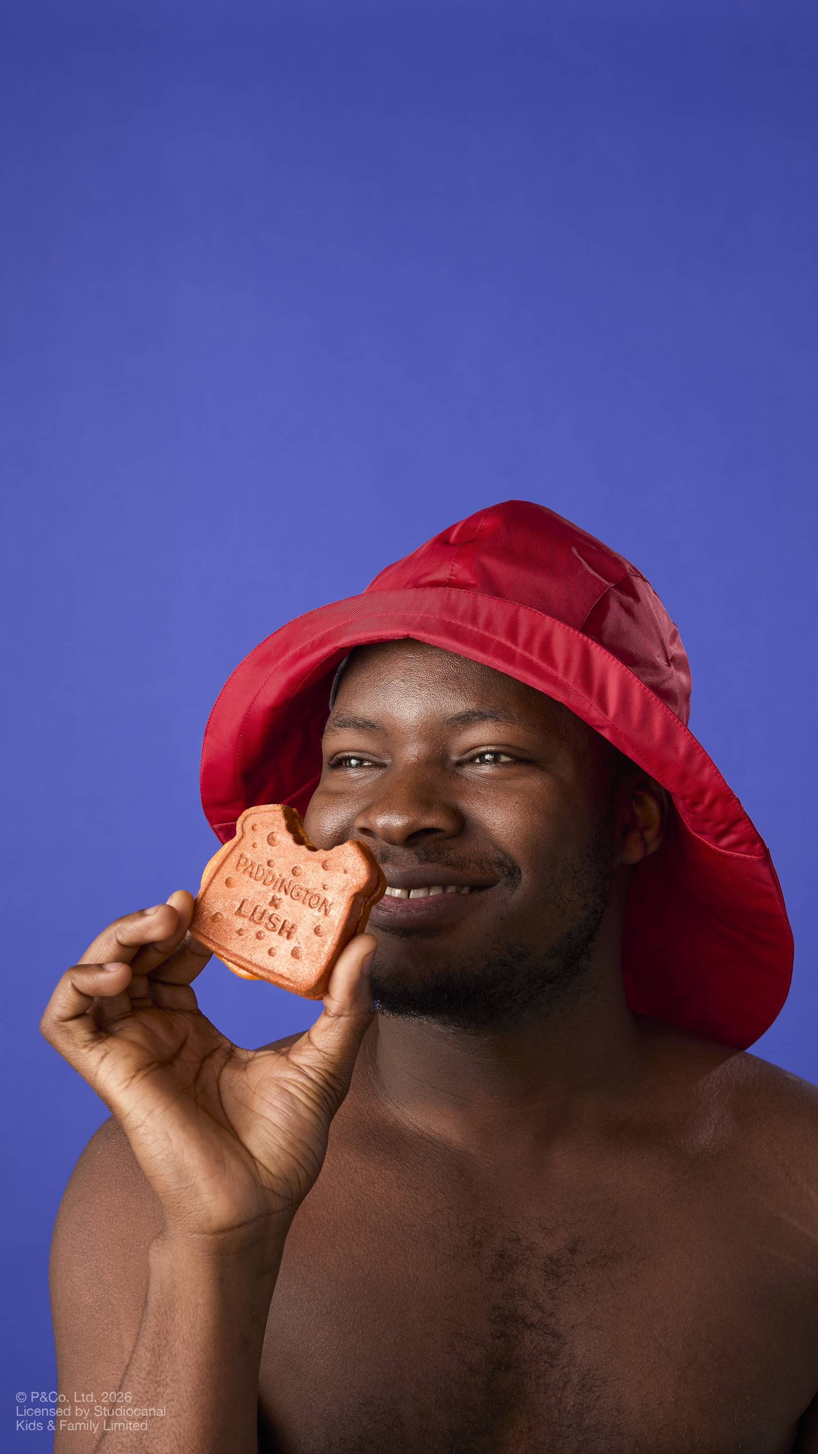 A model in a red rain hat smiles as they hold a "Marmalade Sandwich" bubbleroon bubble bar to their face. The brown, textured bar features a 'bite' and the text 'PADDINGTON x LUSH'. Set against a solid blue background.