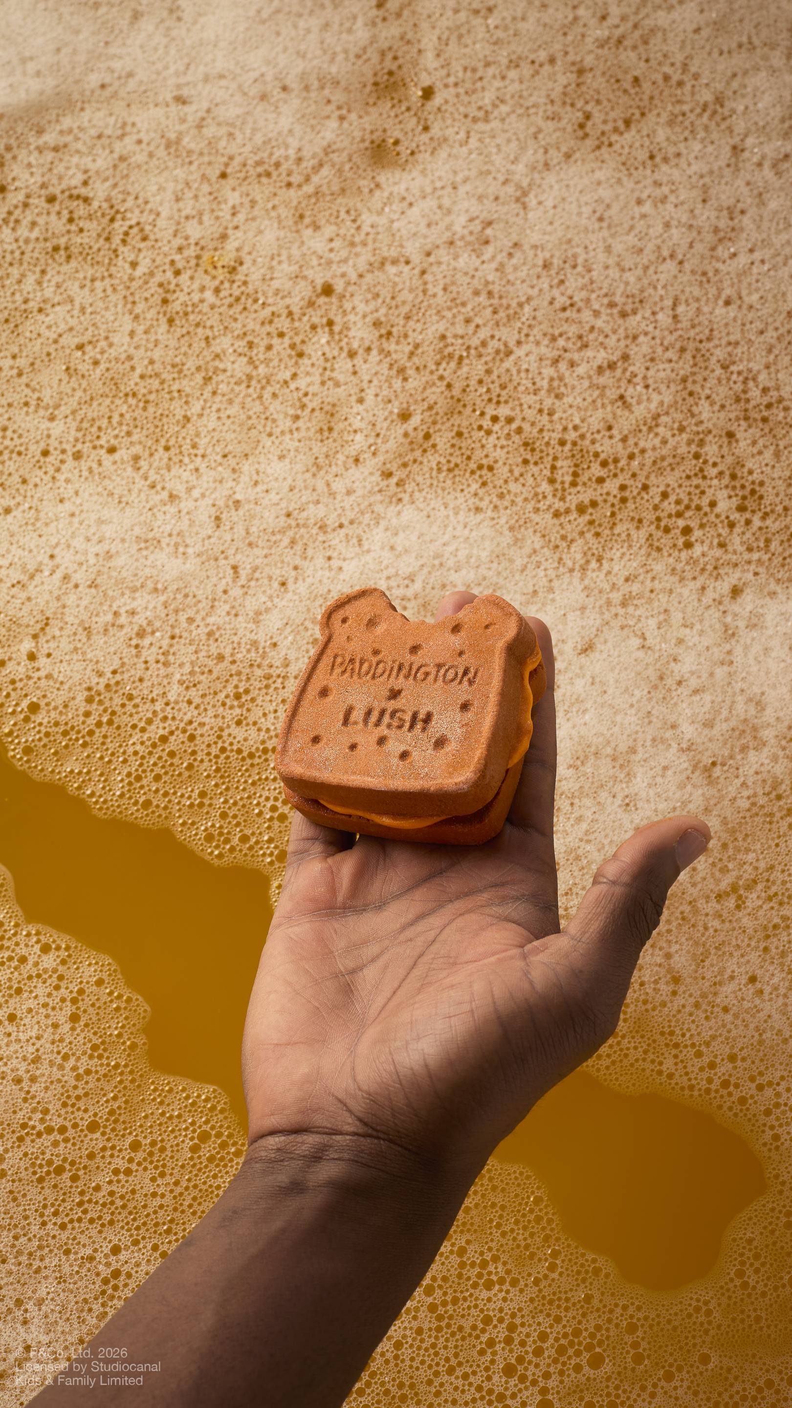 A model's hand holds a "Marmalade Sandwich" bubbleroon bubble bar over golden, foamy bathwater. The brown, textured bubble bar features the embossed text 'PADDINGTON x LUSH' and has a small 'bite' taken out of the top.
