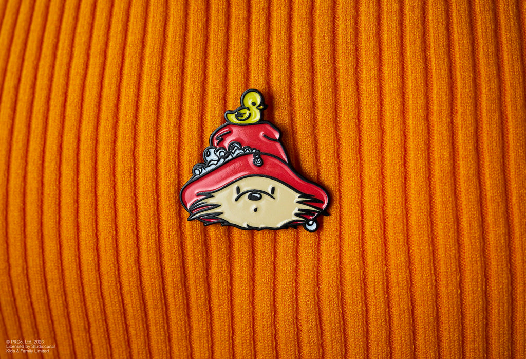 The image shows a Paddington x Lush enamel pin badge pinned to an orange ribbed, corduroy fabric. The badge features Paddington Bear’s head in a red hat with a yellow rubber duck on top and soap bubbles.