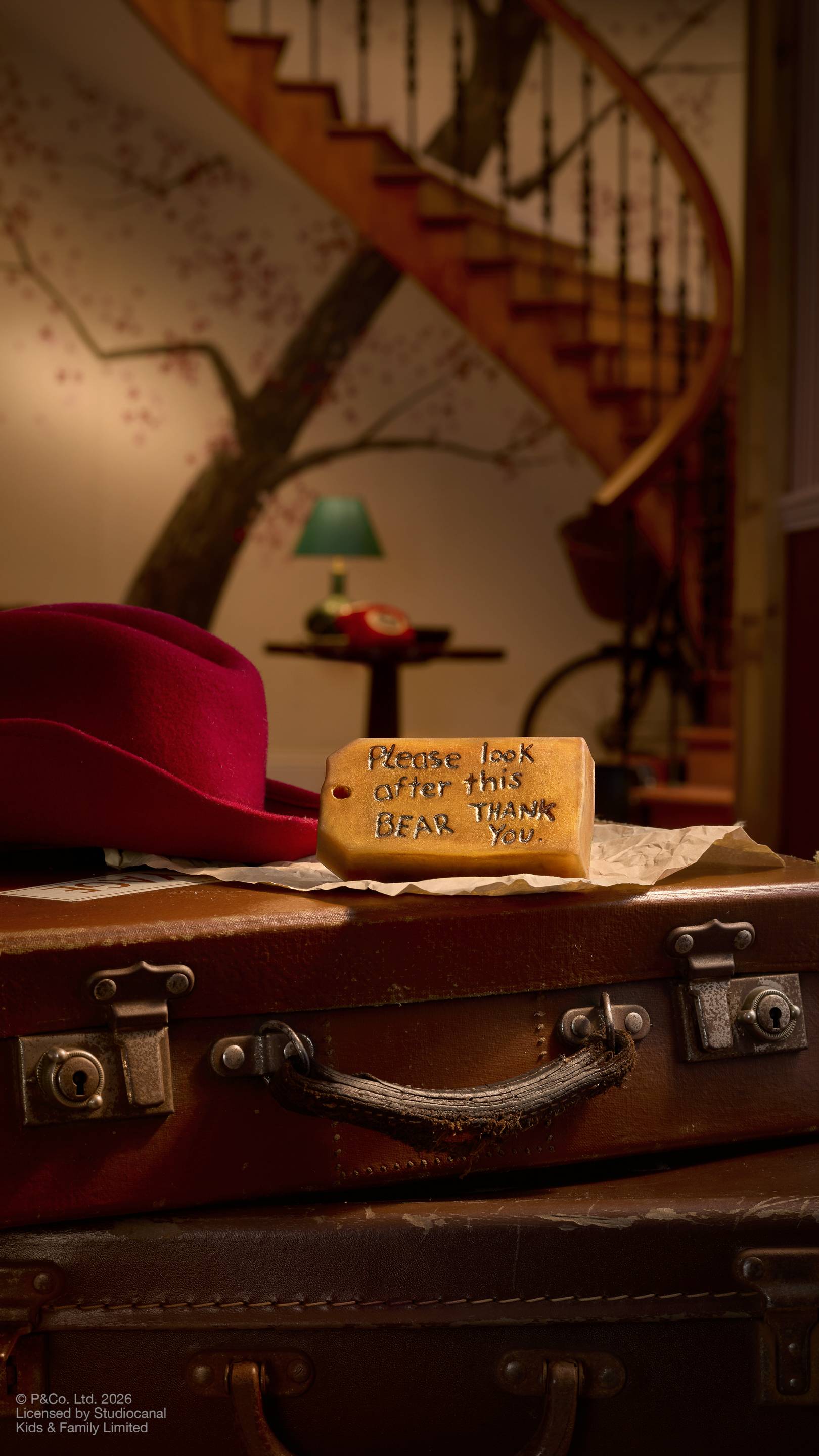 The image shows the brown, luggage-tag-shaped bar of "Please Look After This Bear" soap, inspired by Paddington Bear, sitting on a vintage suitcase next to his red hat. The soap features the embossed message 'Please look after this bear, thank you'.