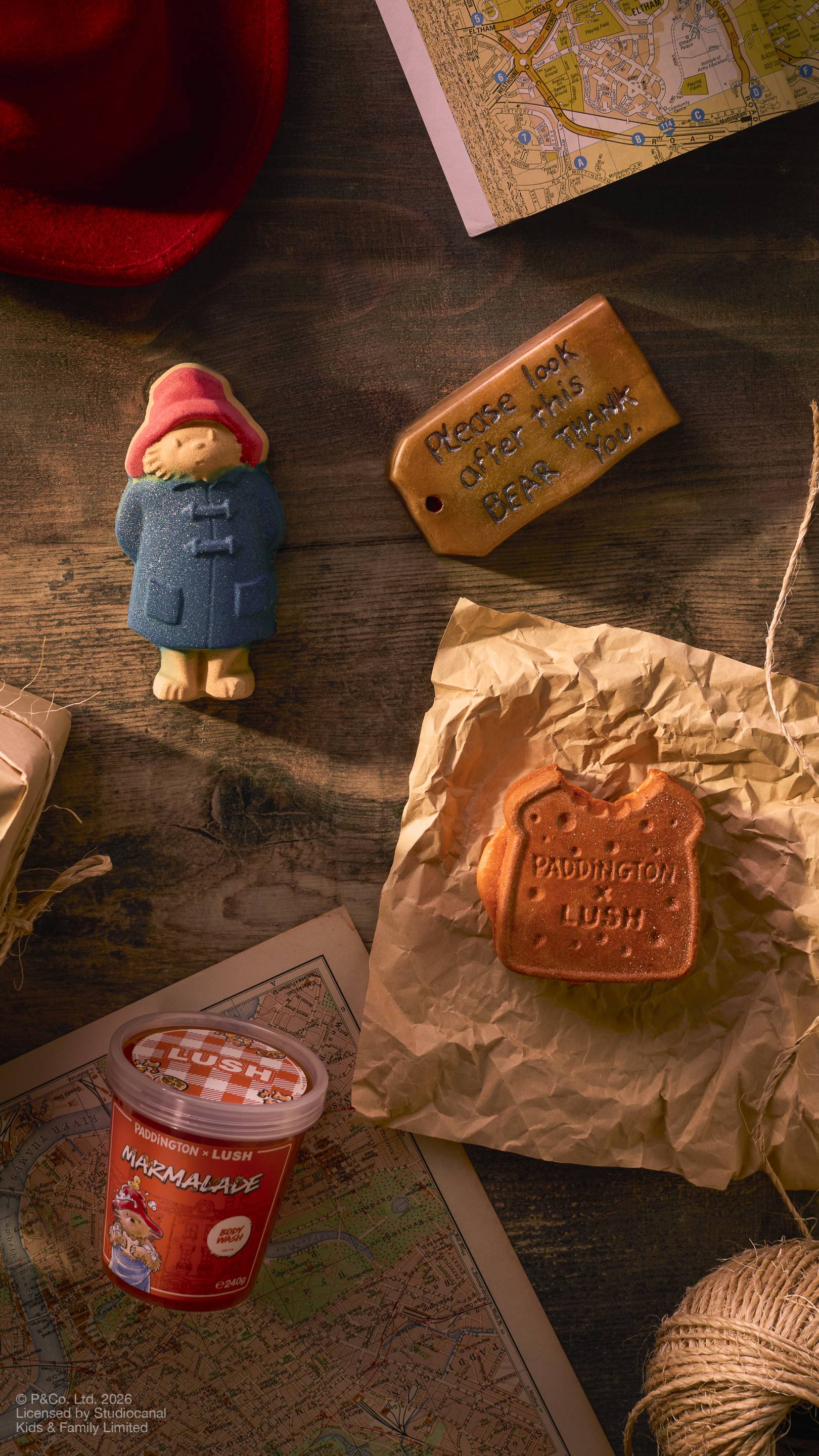 The image shows a collection of Paddington Bear-themed Lush products on a dark wooden surface, including a Paddington bath bomb, a marmalade sandwich-shaped bubble bar, and a 'Marmalade' body wash, around Maps, string, and a red hat.
