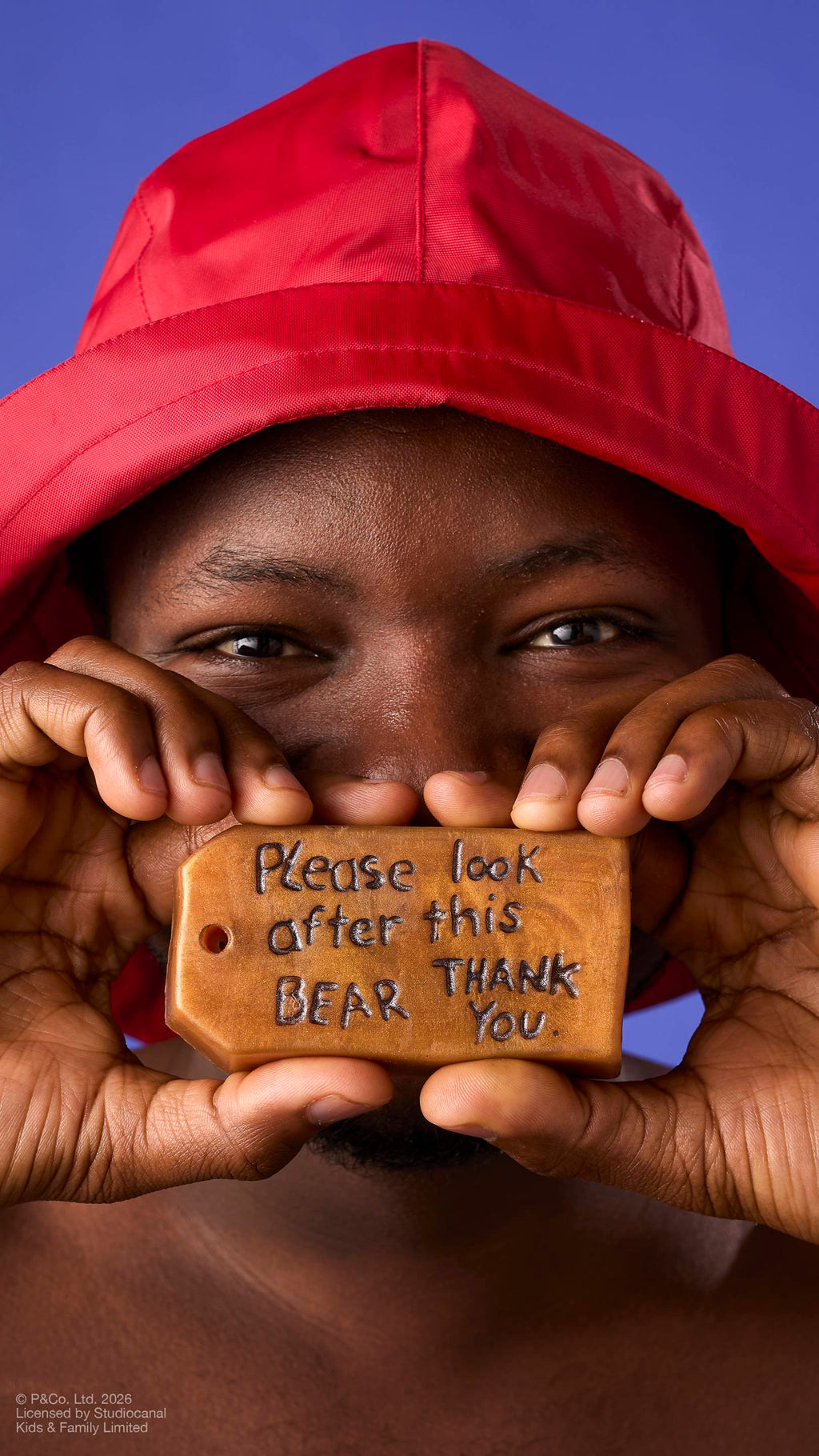 A model wearing a red rain hat holds a brown, luggage-tag-shaped soap in front of their face. The soap features the embossed, handwritten-style message 'Please look after this bear, thank you'. On a solid blue background.