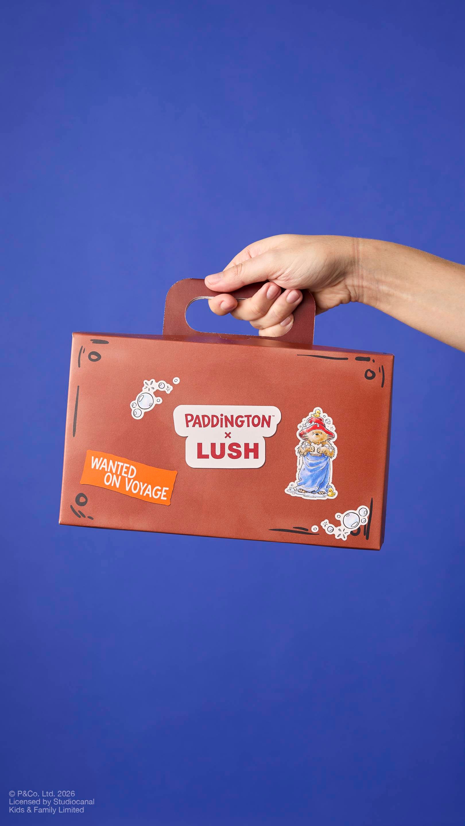 A brown cardboard gift box designed to look like a small suitcase, held by hand by its handle. It features ‘PADDINGTON x LUSH’ branding, an illustration of the bear in a towel, and a ‘WANTED ON VOYAGE’ sticker against a blue background.