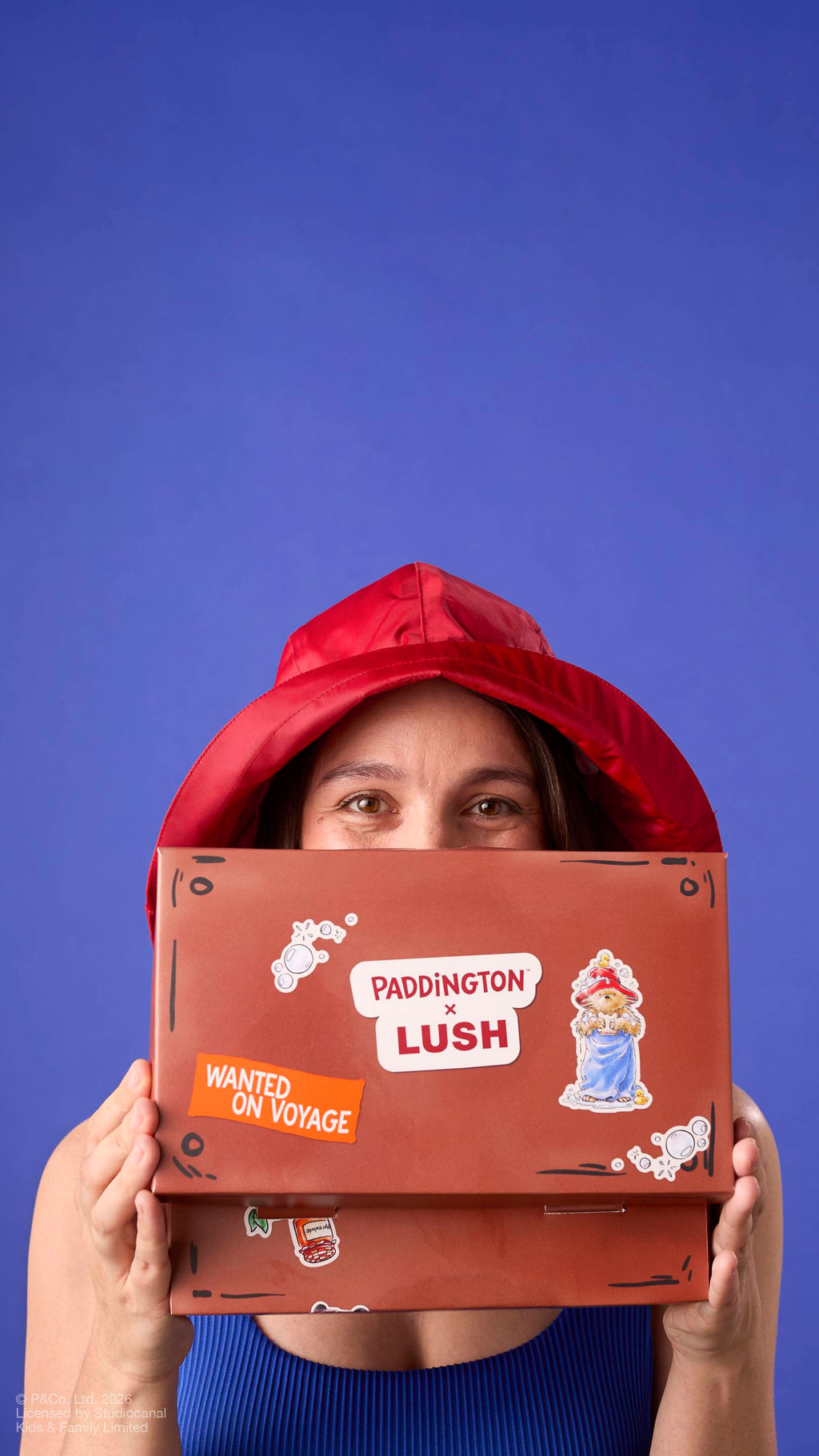 A model wearing a red rain hat peeks over a brown, suitcase-shaped gift box. The packaging features a 'PADDINGTON x LUSH' logo, a 'WANTED ON VOYAGE' sticker, and an illustration of the bear on a plain blue background.