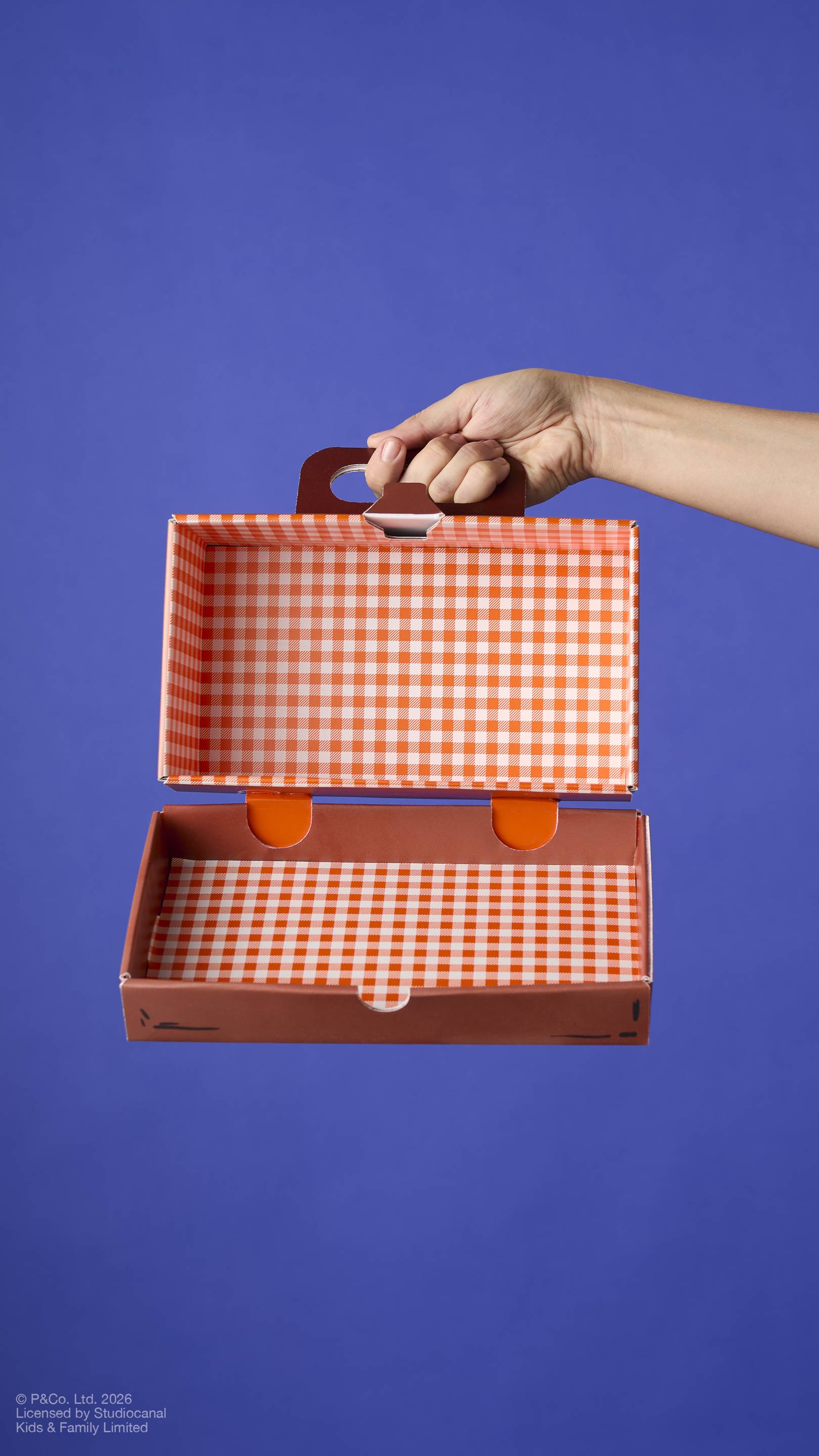 An open cardboard gift box shaped like a suitcase, held by a hand. The interior is lined with a bright orange and white gingham pattern. Set against a solid blue background.