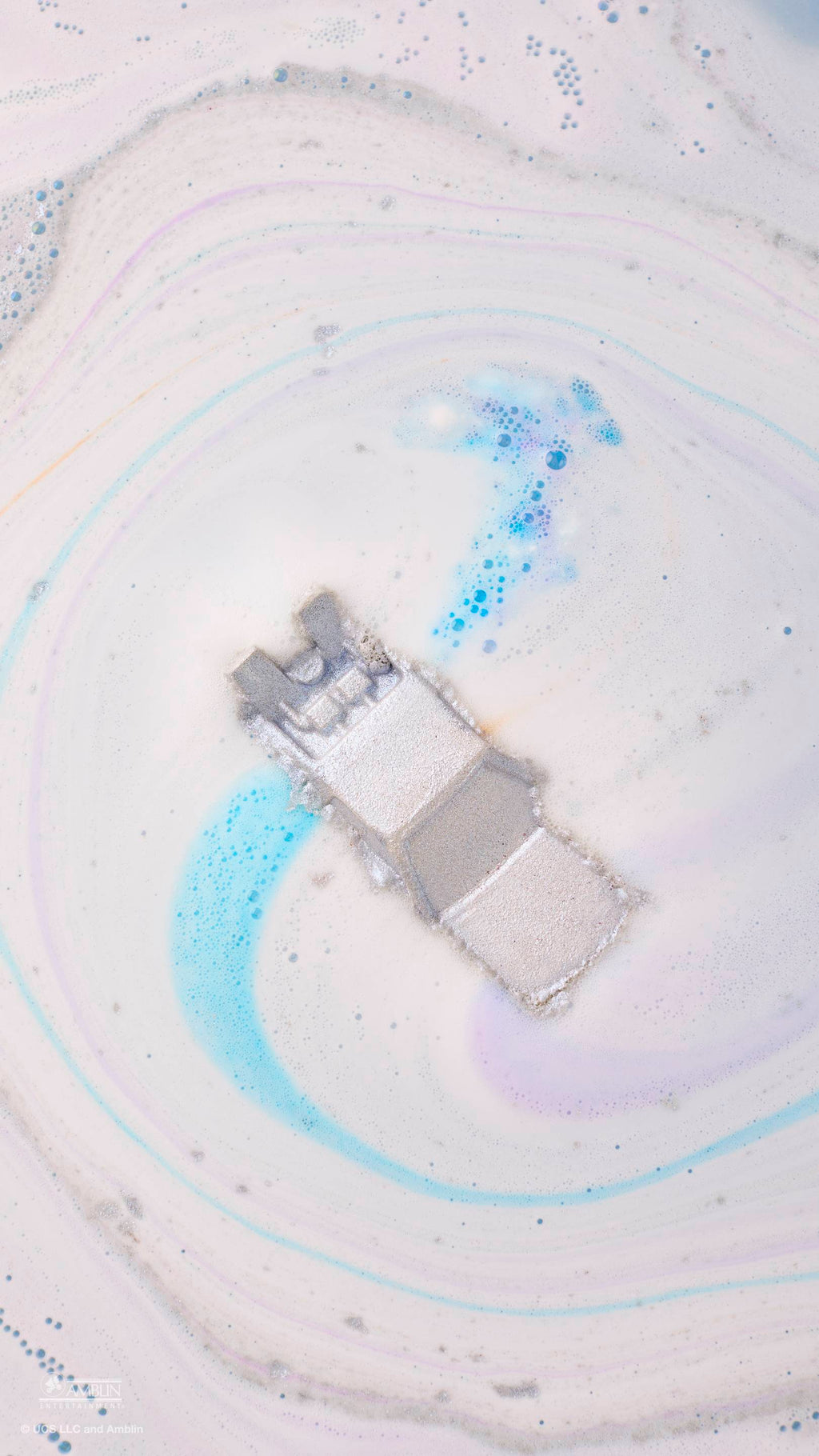 An aerial view of a silver, DeLorean car-shaped bath bomb dissolving in water. It creates a swirling pattern of white, pale purple, and bright blue foam, with the metallic car leaving a shimmering trail as it floats in the centre of the tub.
