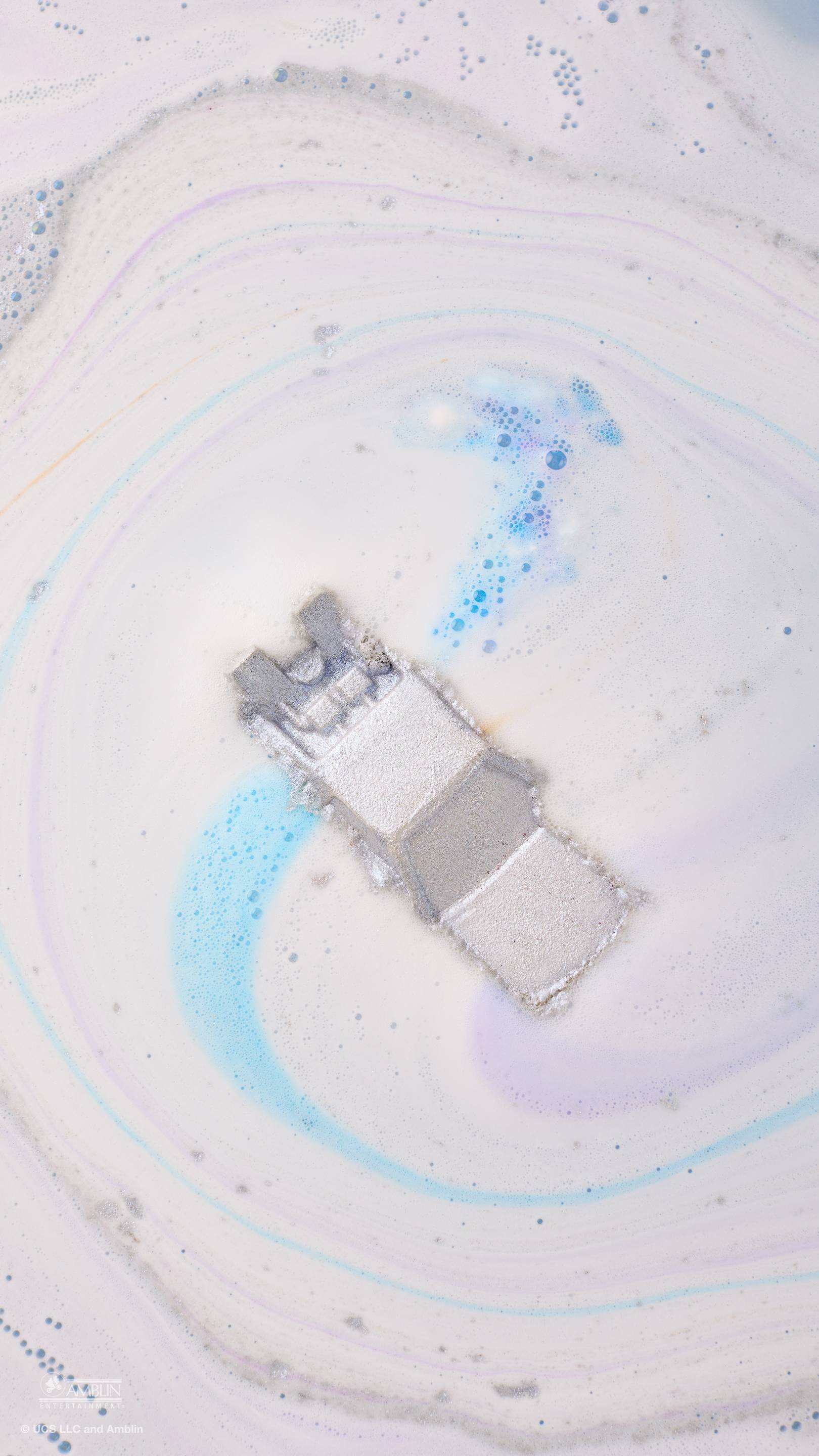 An aerial view of a silver, DeLorean car-shaped bath bomb dissolving in water. It creates a swirling pattern of white, pale purple, and bright blue foam, with the metallic car leaving a shimmering trail as it floats in the centre of the tub.