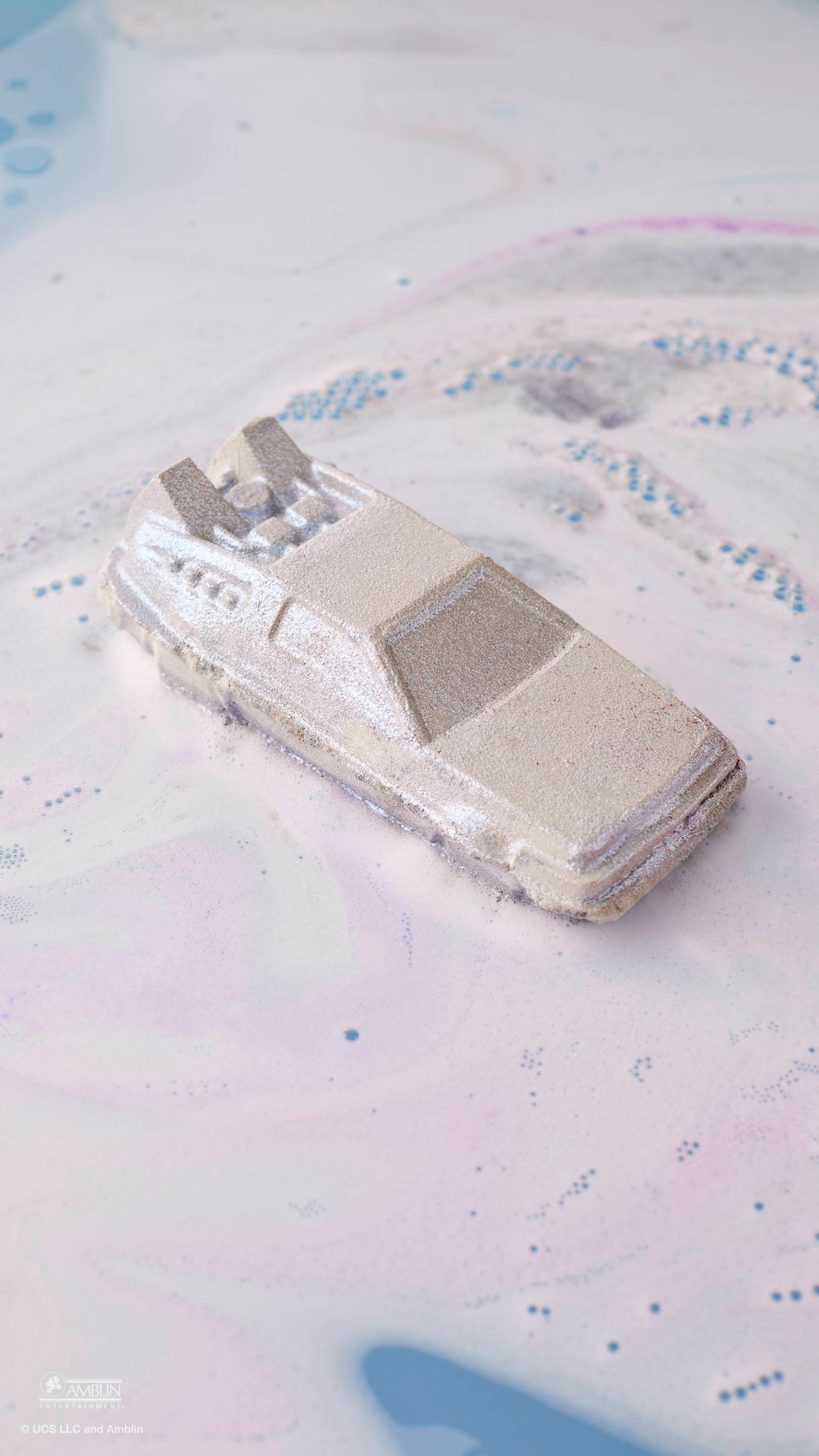 A silver, car-shaped bath bomb floats in bathwater, creating swirling clouds of white and pale purple foam. The metallic, angular car is inspired by a DeLorean and leaves a shimmering trail as it dissolves across the surface of the water.