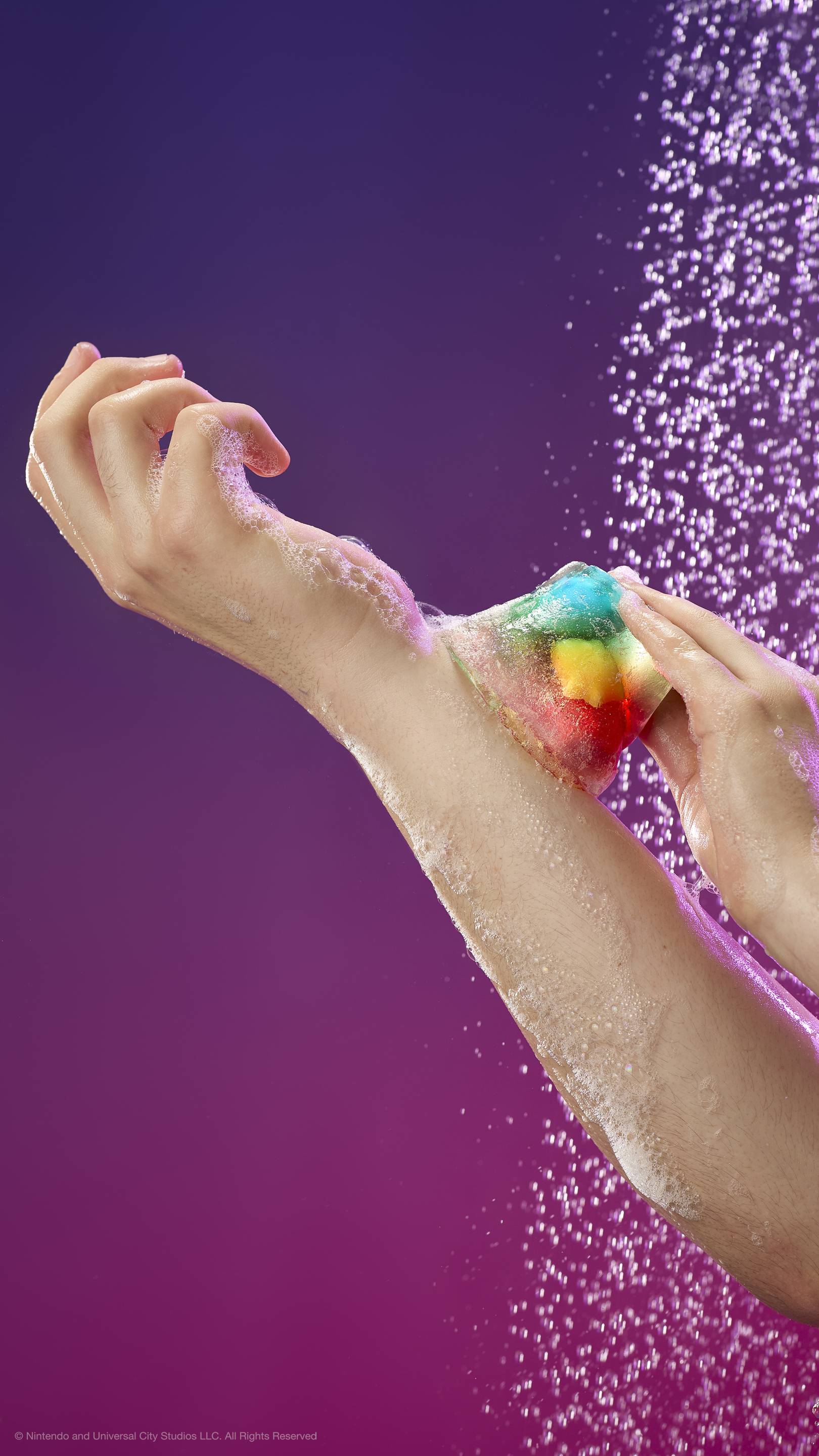 Hand glides translucent shower jelly with colorful star-shaped Lumas over a soapy arm under a shower spray. Set against a purple-pink gradient background. Includes plastic-free glitter.