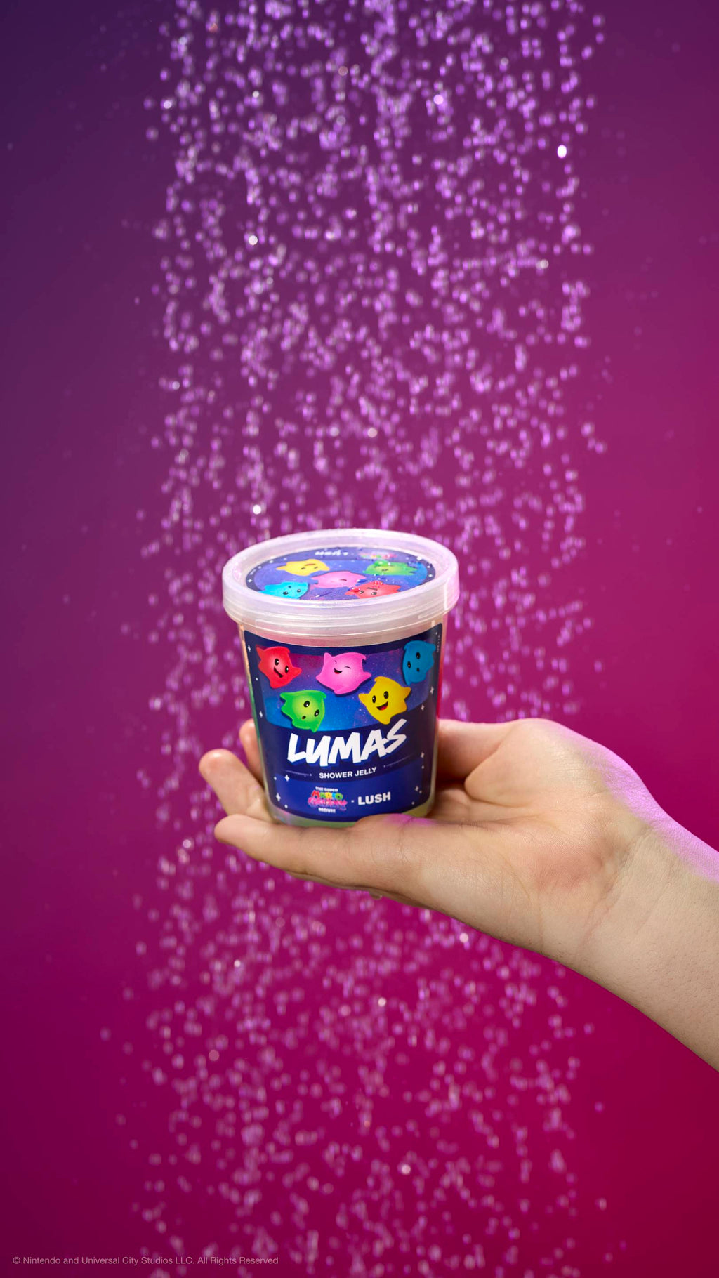 Hand holds a pot of Lumas shower jelly under a shower spray against a purple-pink gradient. Label features colorful Luma characters and movie logos. Includes plastic-free glitter.