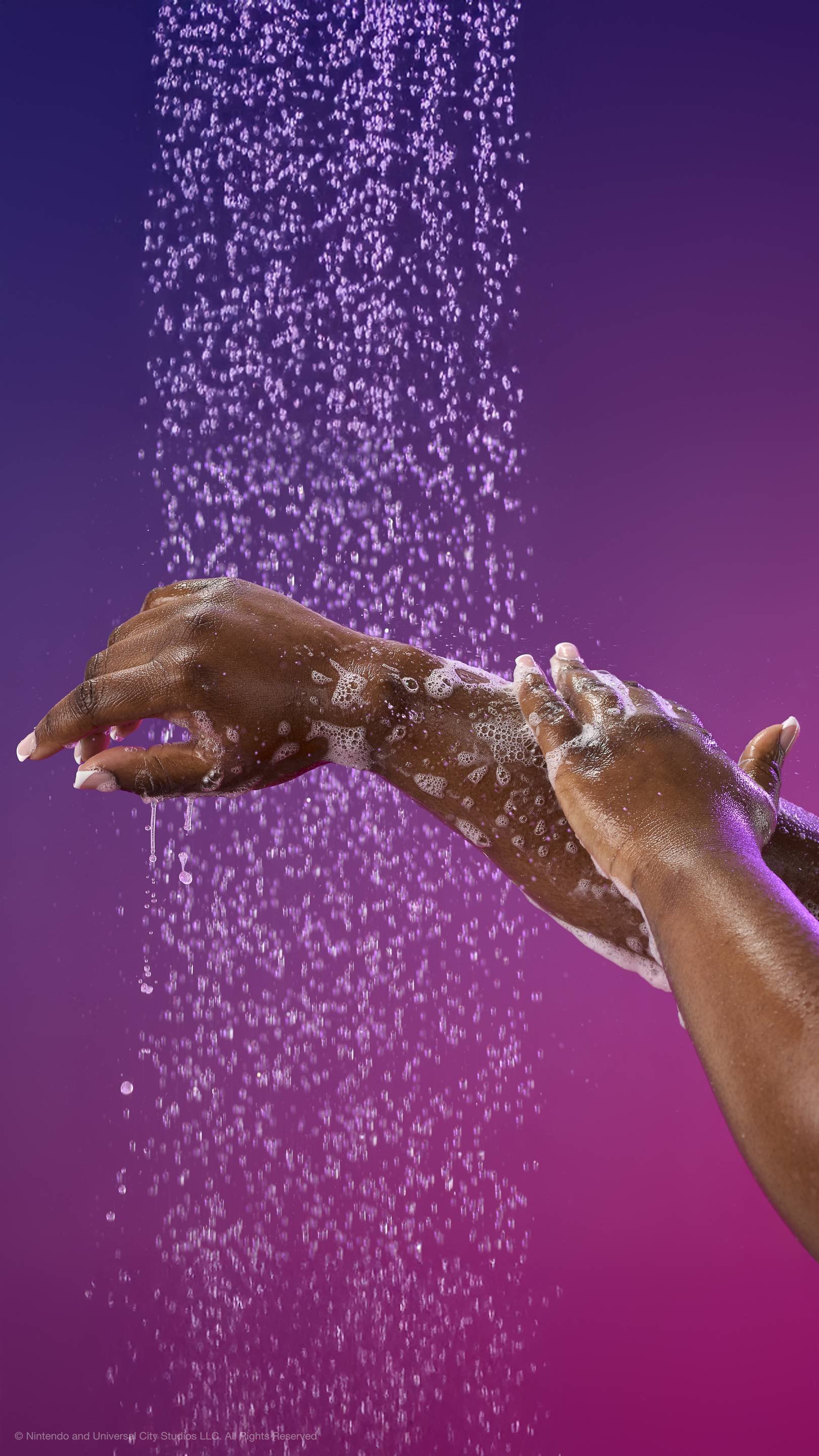 Hands lather an arm with white foam under a shower spray against a purple-magenta gradient. Features many fine water droplets and white suds across the skin.