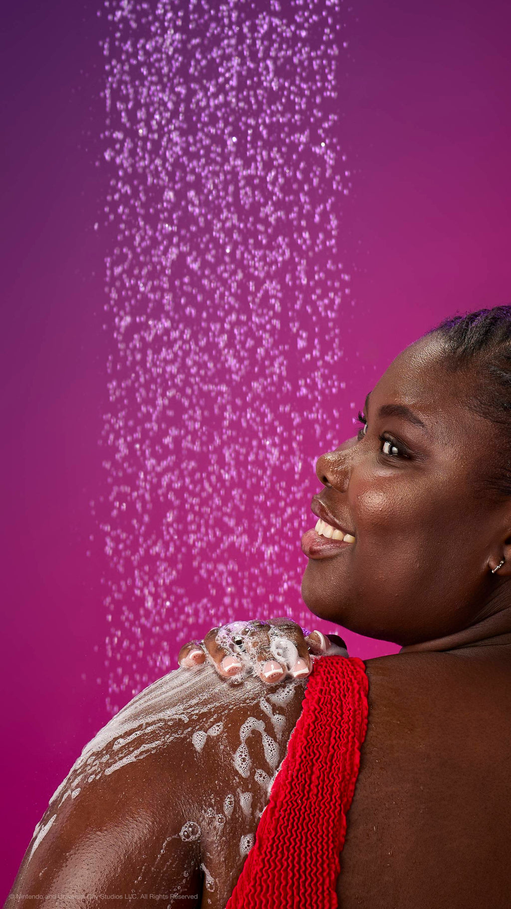 A woman in a red top smiles over her shoulder while lathering her shoulder with white foam under a shower spray. Against a purple-magenta gradient background with many fine water droplets.