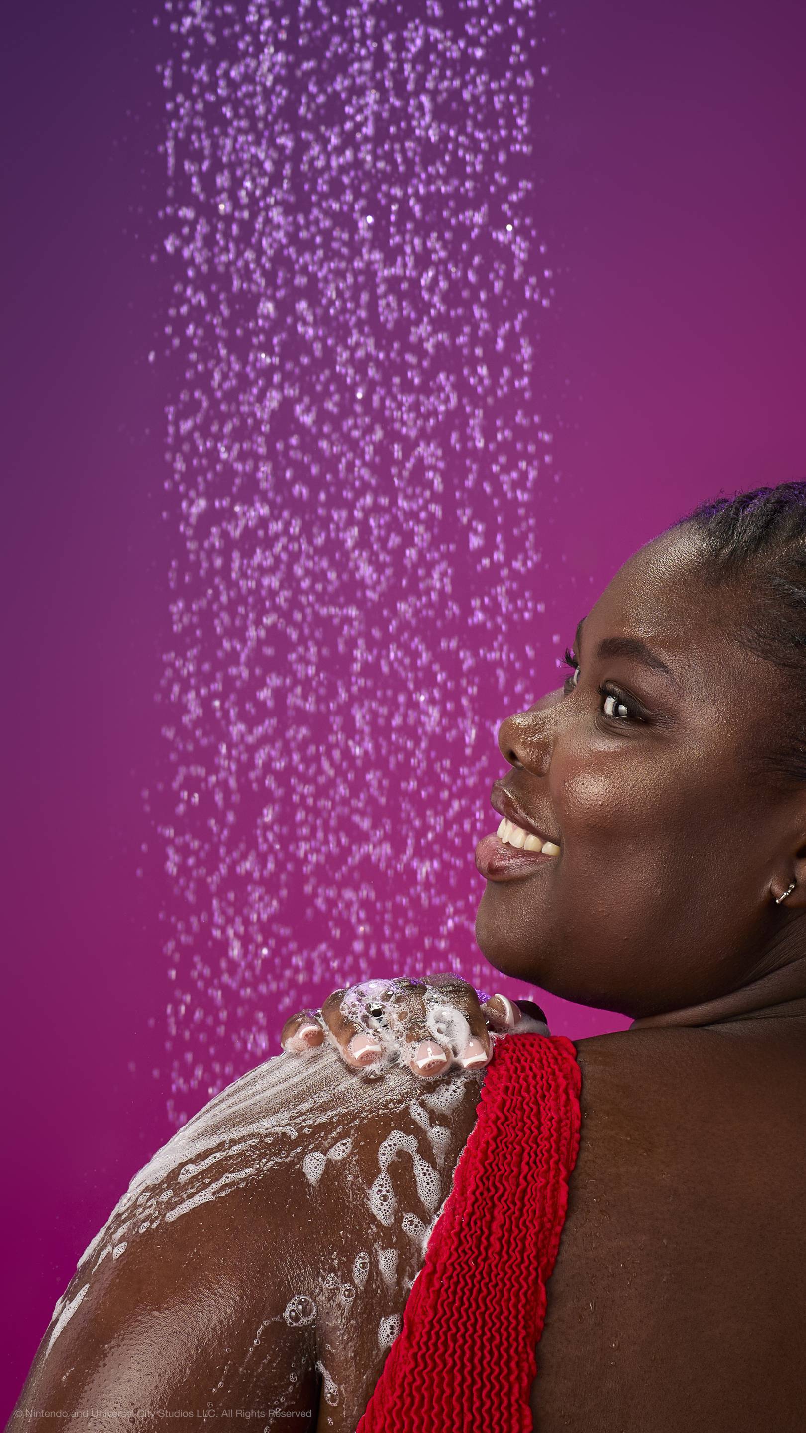 A woman in a red top smiles over her shoulder while lathering her shoulder with white foam under a shower spray. Against a purple-magenta gradient background with many fine water droplets.