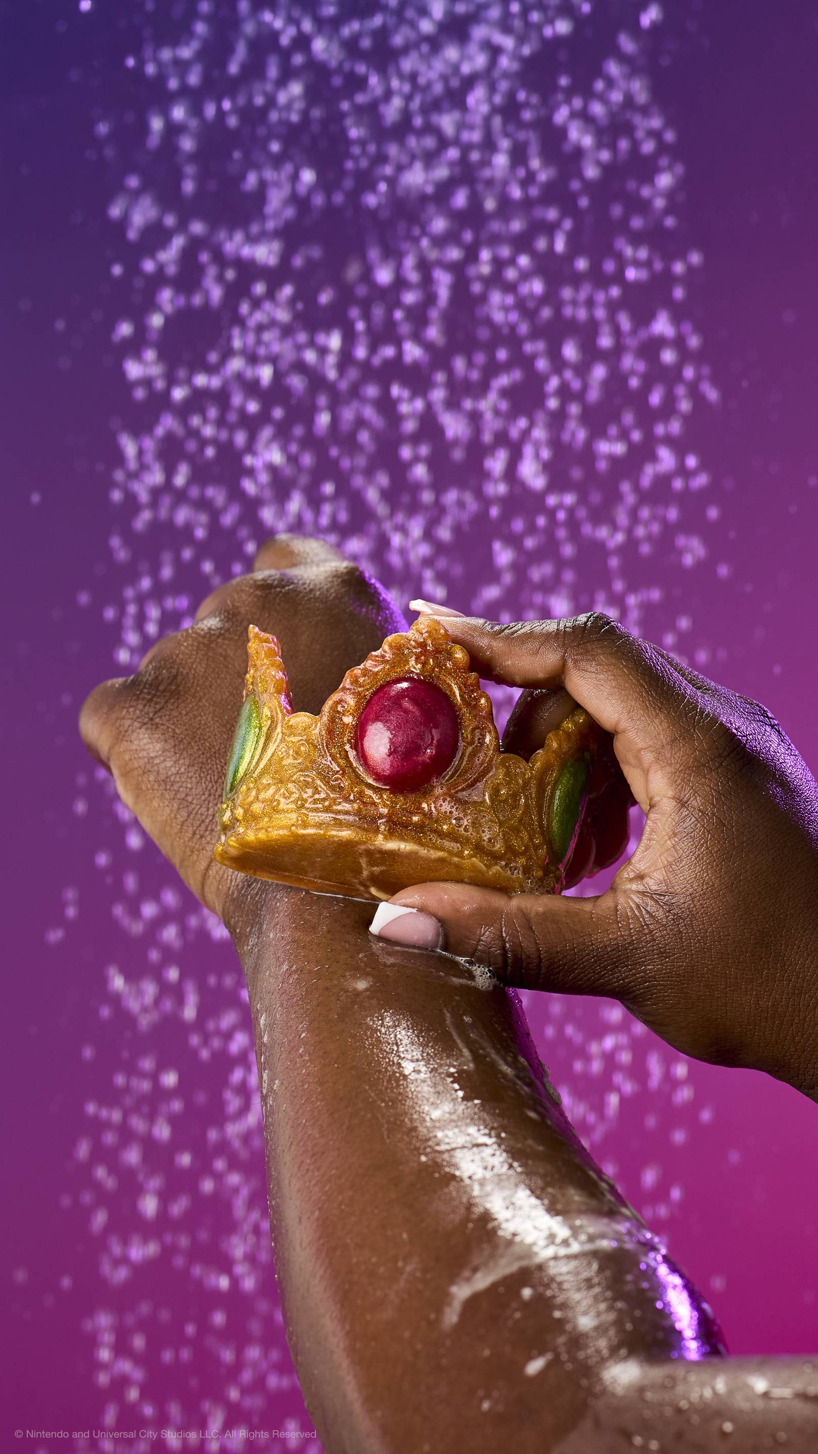 A hand glides the shimmering gold Princess Peach crown soap over a soapy forearm under a shower spray. Features red and green jewels against a purple-pink gradient background.