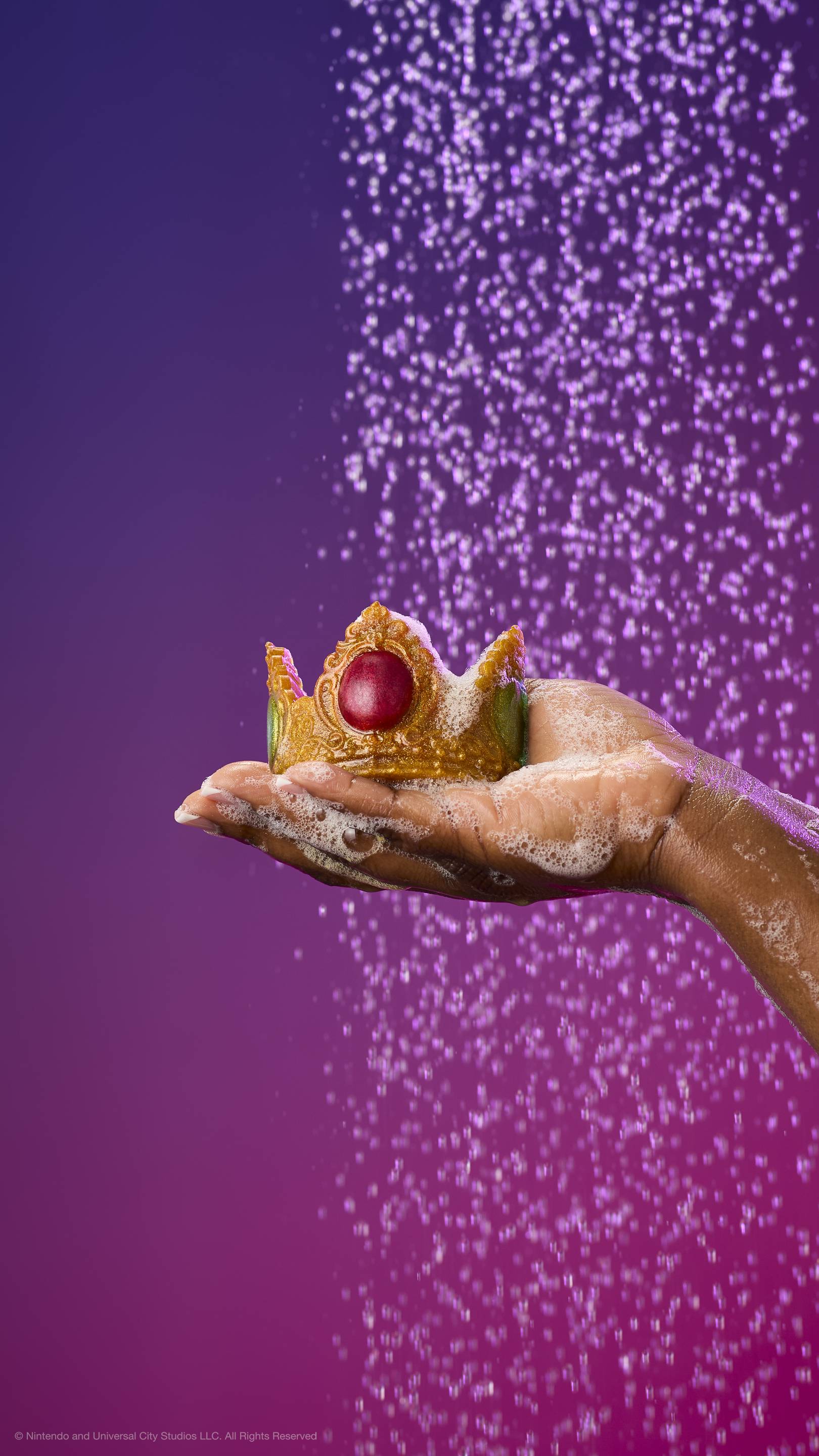 Shimmering gold Princess Peach crown soap with a large red jewel held in a soapy palm under a shower spray. Against a purple-magenta gradient with plastic-free glitter.
