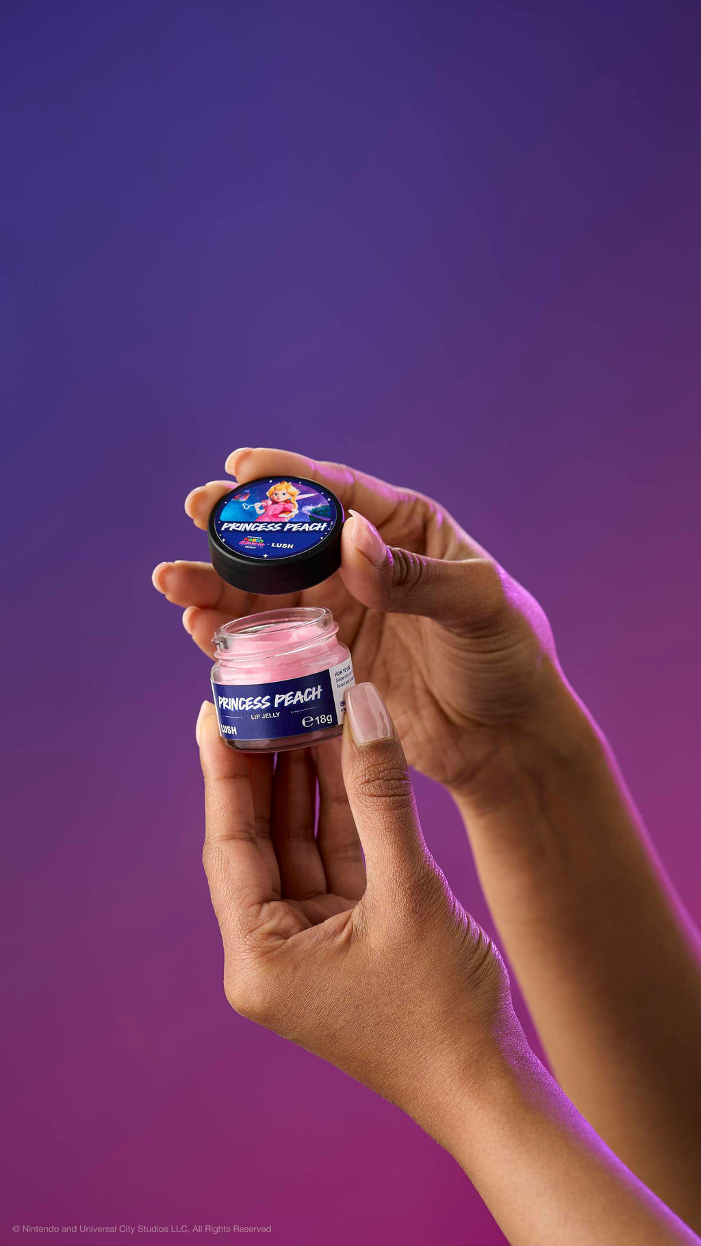 Two hands hold an open pot of creamy pink Princess Peach lip jelly and its lid against a purple-magenta gradient. The lid features Princess Peach holding a heart-shaped wand.