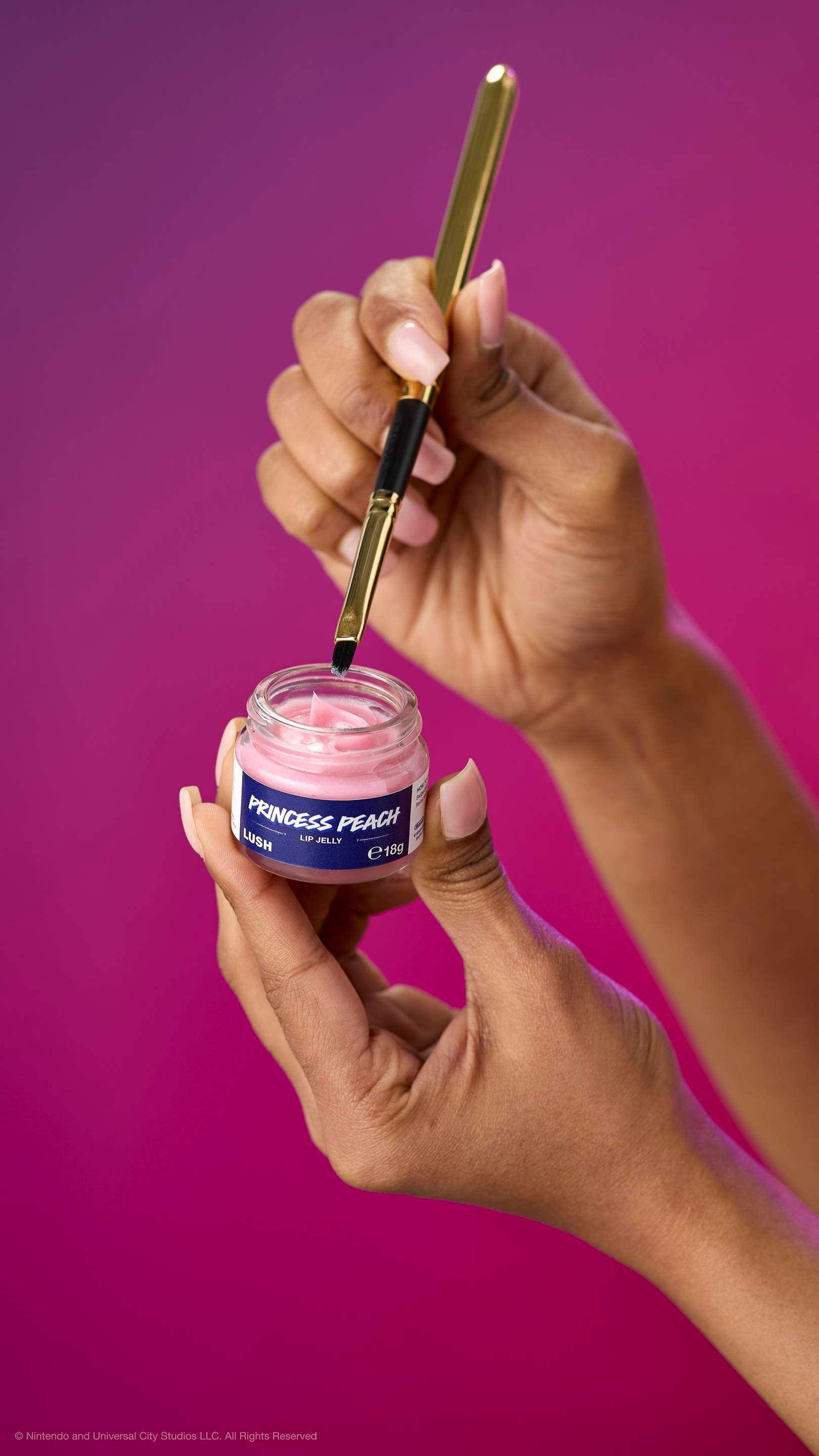 Hands hold an open pot of creamy pink Princess Peach lip jelly while a gold lip brush picks up product. Against a purple-magenta gradient background.