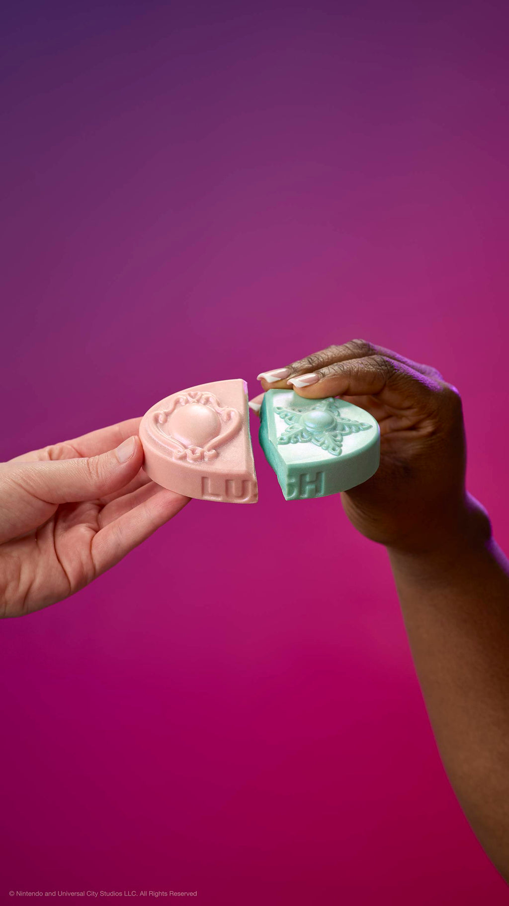 Two hands join the pink and teal halves of the Princess Power body balm against a purple-pink gradient. The pieces feature embossed brooch and star designs and the LUSH logo on the side.