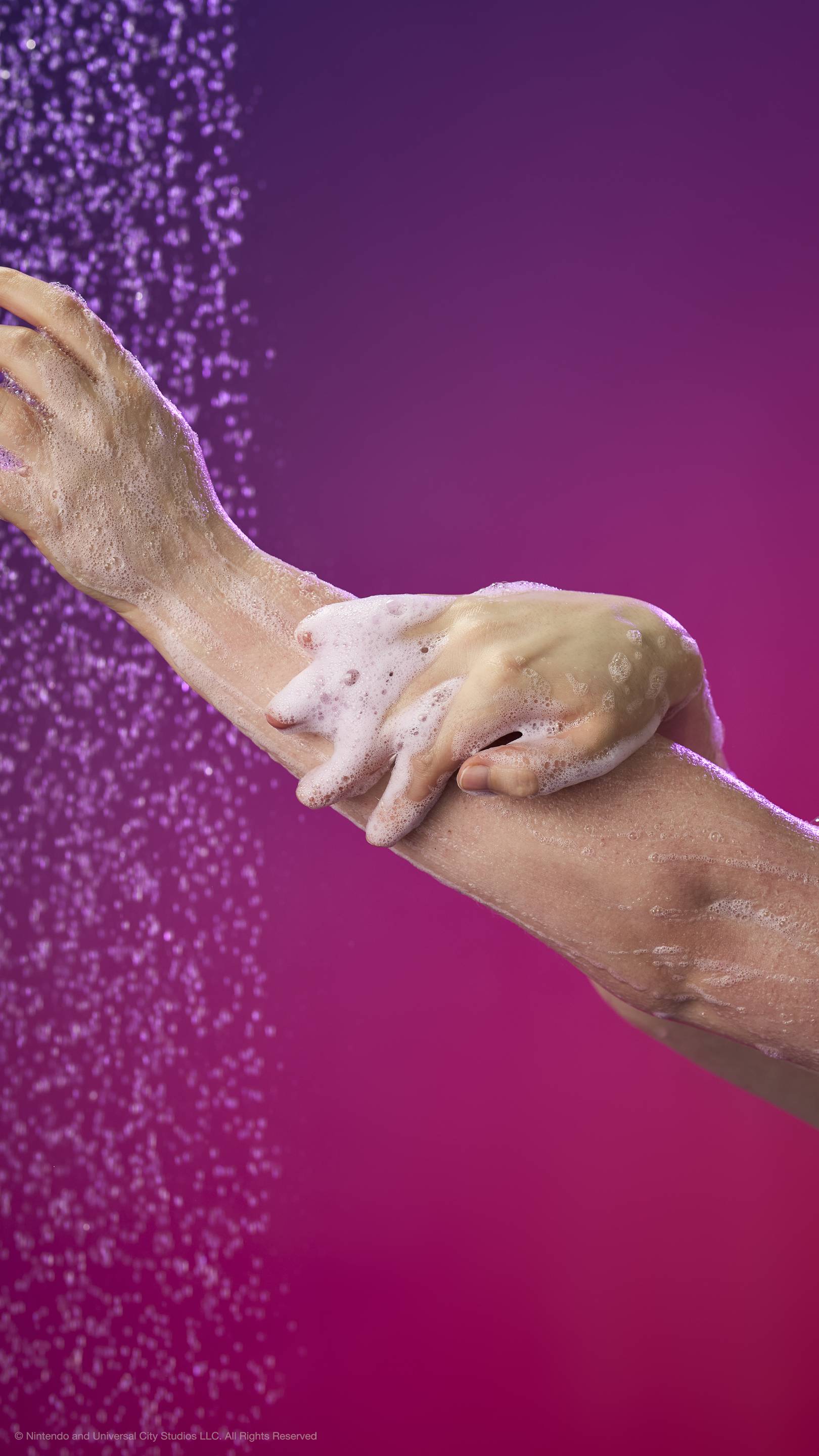 Hands lather white foam onto an arm under a shower spray against a purple and pink gradient background.
