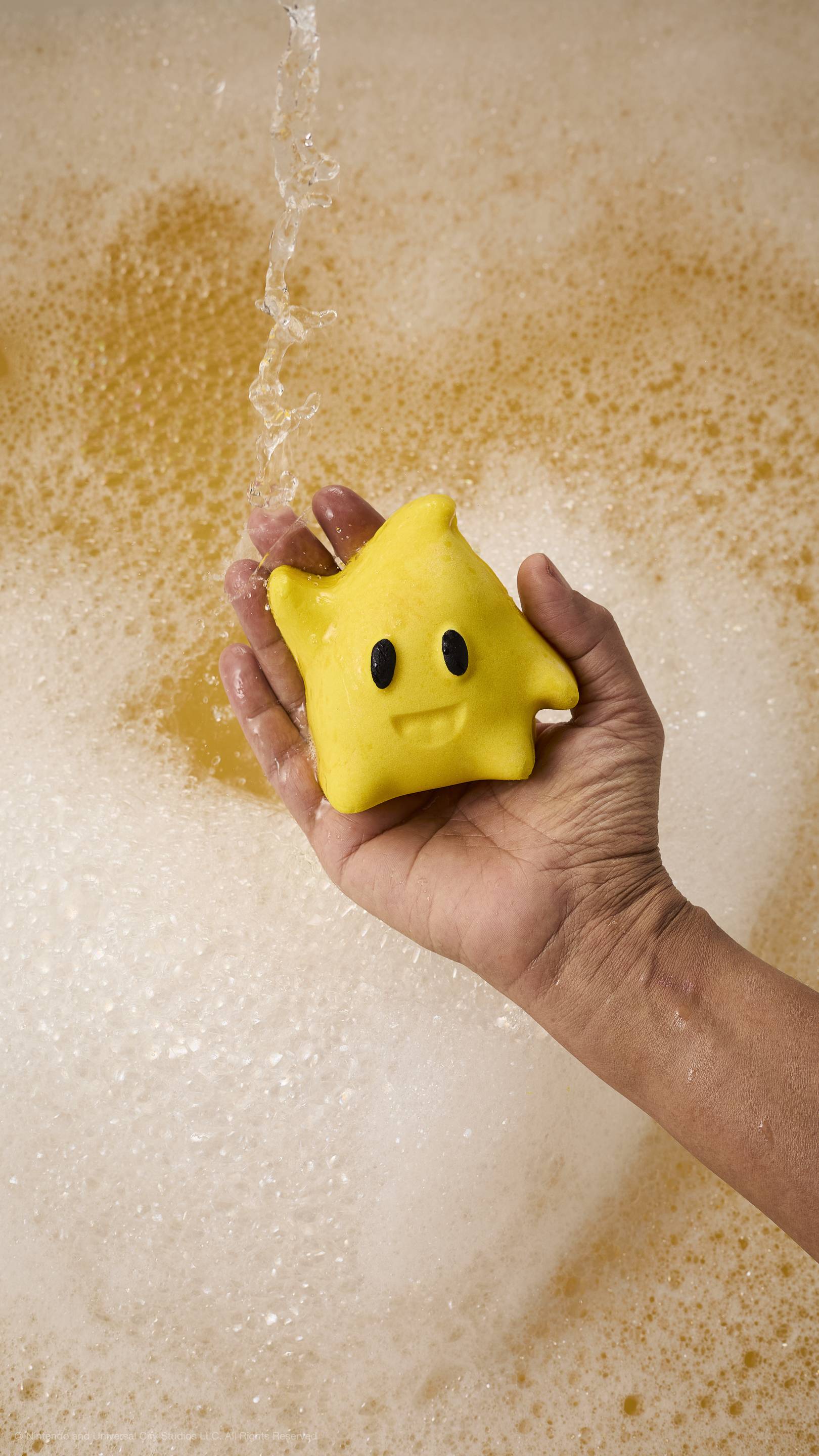 A hand holds a yellow star-shaped Luma bubble bar with black eyes and a smile under running water. The bathtub is filled with thick white foam and golden-toned bubbles.