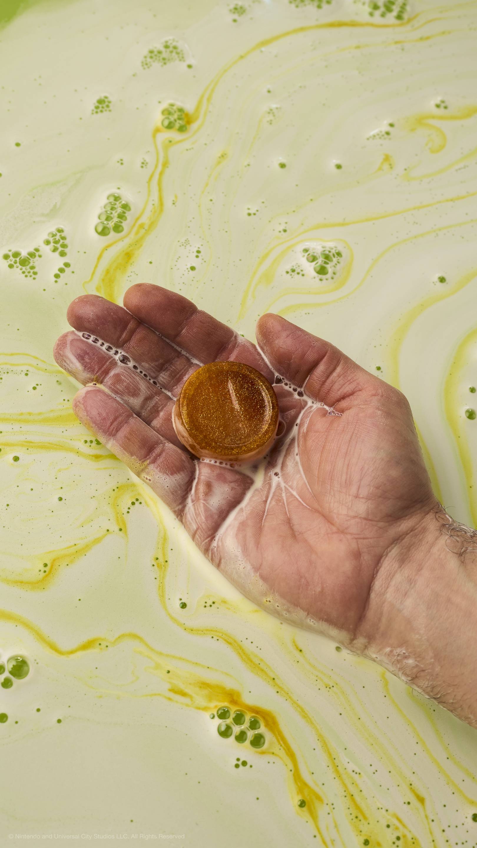 A hand holds a small, gold-colored circular soap shaped like a Mario coin over light-yellow bathwater with white foam.