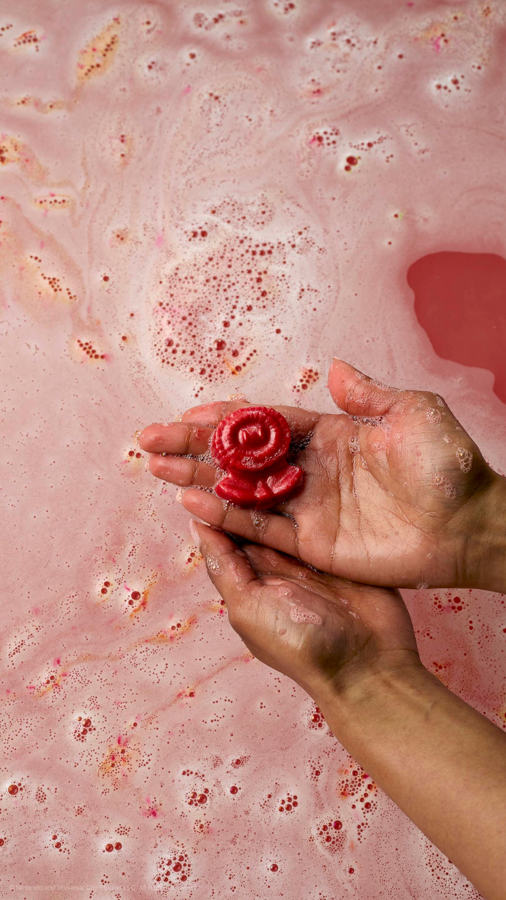 Hands hold a small, red soap shaped like a Fire Flower over bubbly, pink-toned bathwater.