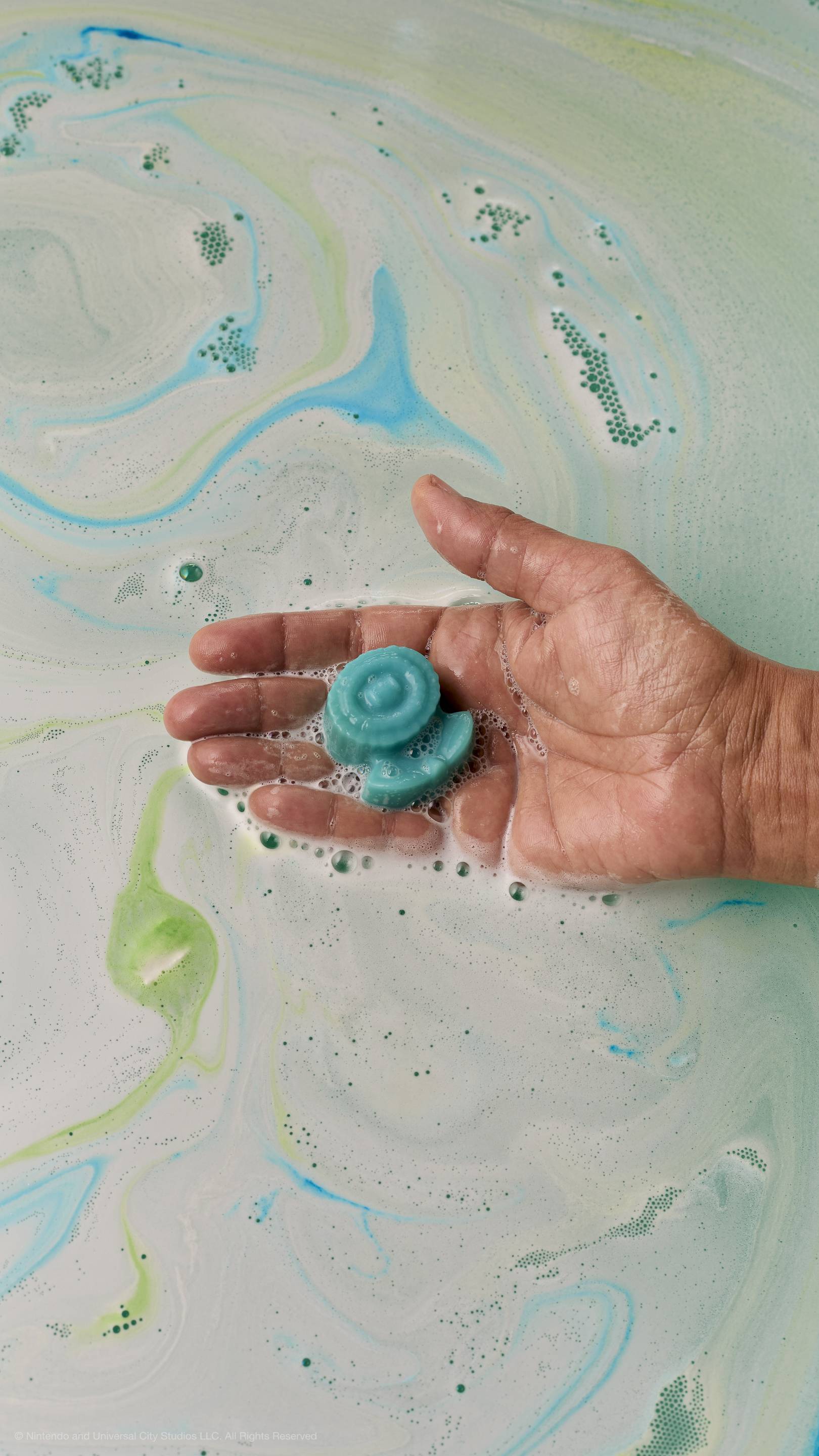 A hand holds a small, light-blue soap shaped like an Ice Flower over bathwater filled with swirls of white, light-blue, and green foam.