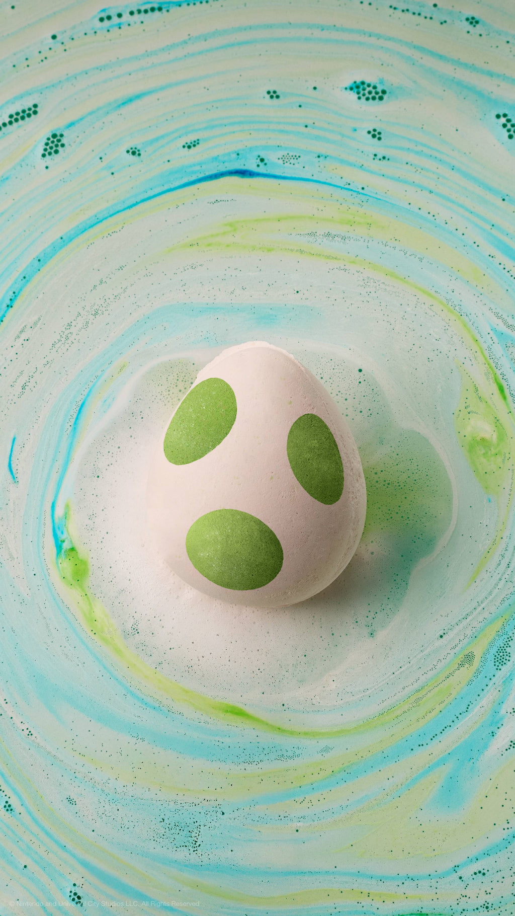 A white Yoshi egg bath bomb with green spots floats in water, surrounded by swirling patterns of green, blue, and white foam.
