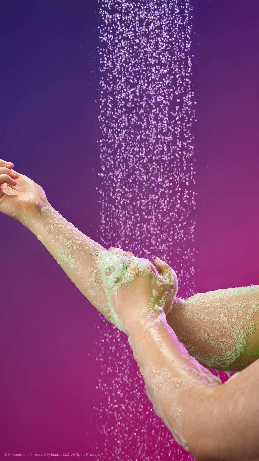 Hands lather an arm with pale green foam under a shower spray against a purple-magenta gradient. Fine water droplets fall through the air around the soapy skin.