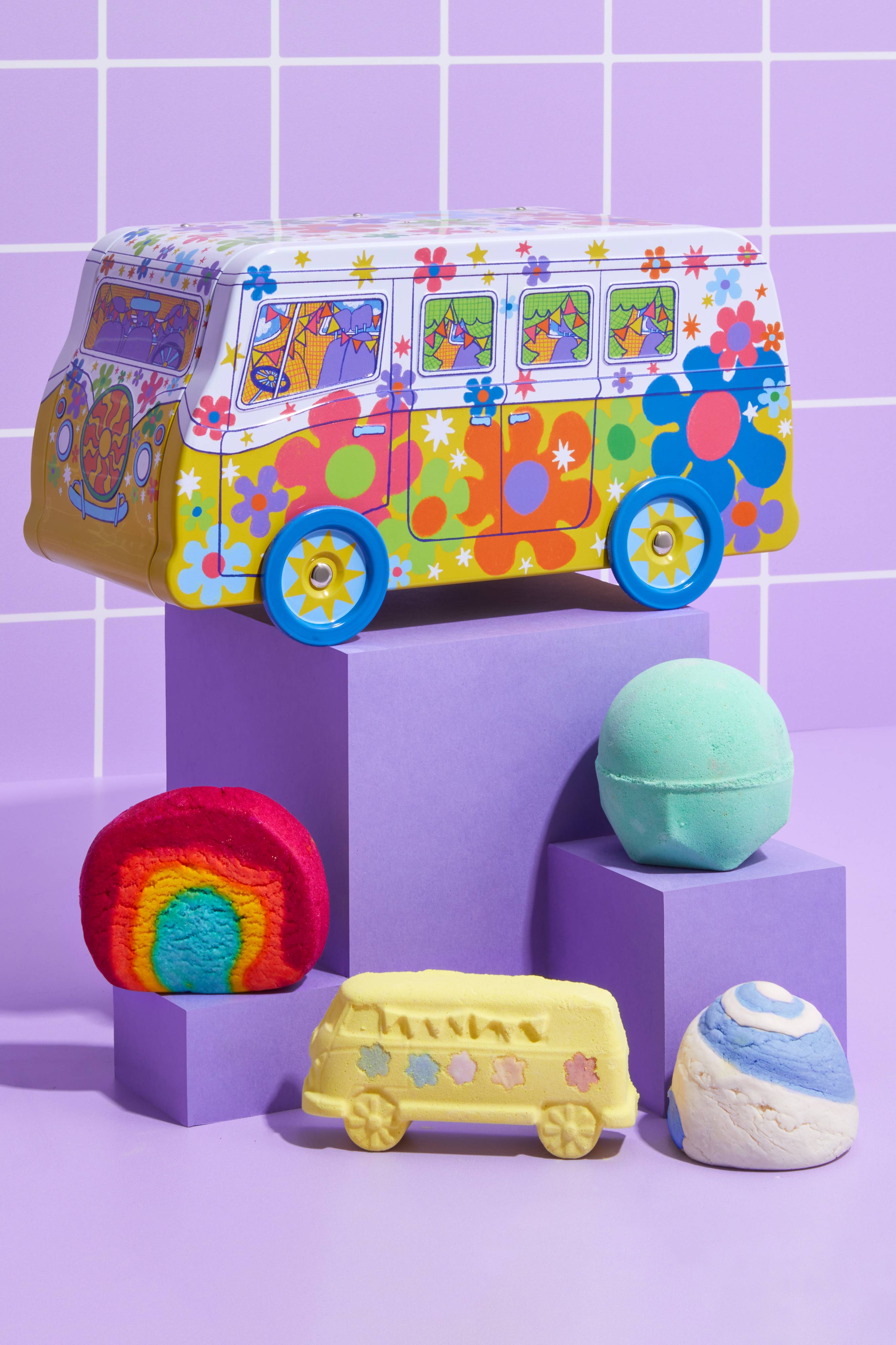 The image shows a colourful psychedelic 'Magic Bus' camper van tin sitting on a purple plinth. Four bathing products are arranged on steps, against a purple tiled wall with white grout, including two bath bombs and two bubble bars.