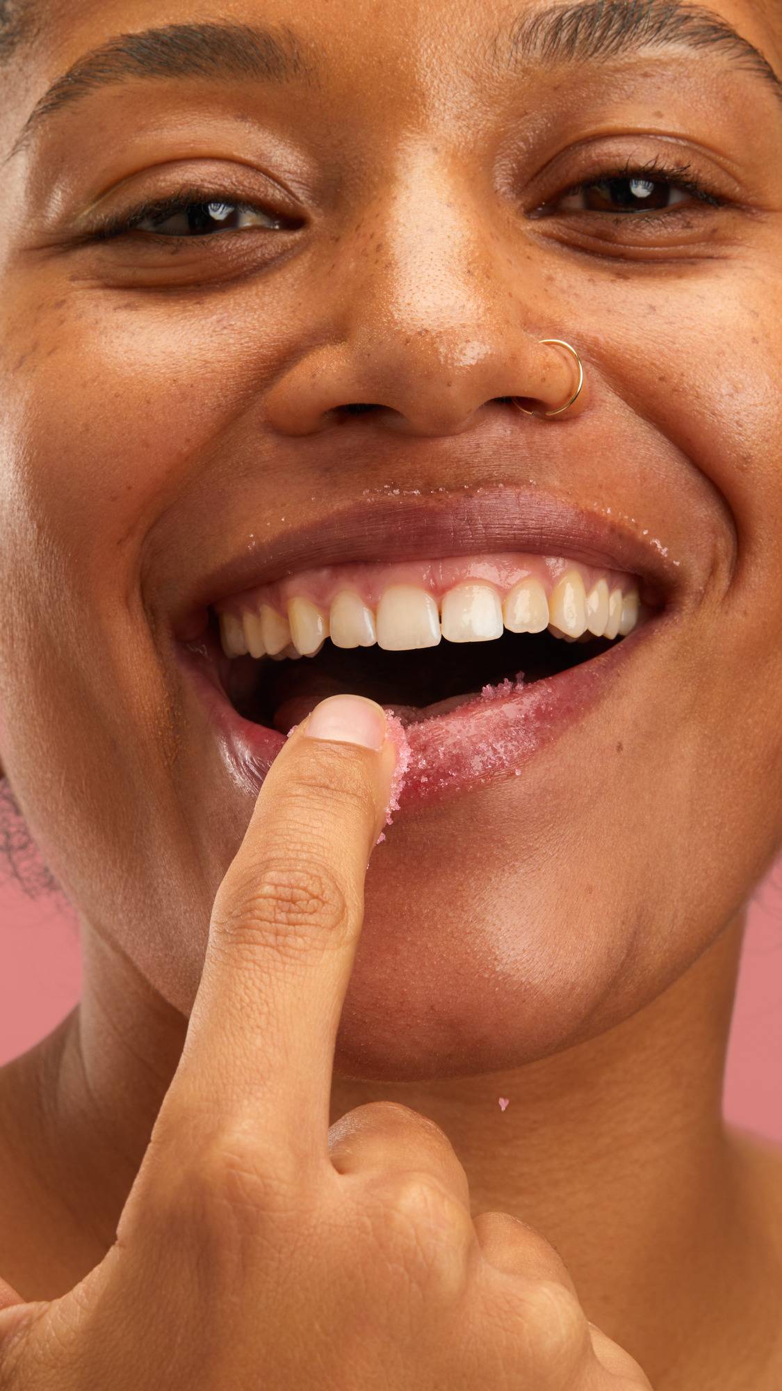 A close-up shot of a model smiling widely while applying the Lush "Bubblegum" lip scrub to their bottom lip with a finger against a plain pink background.