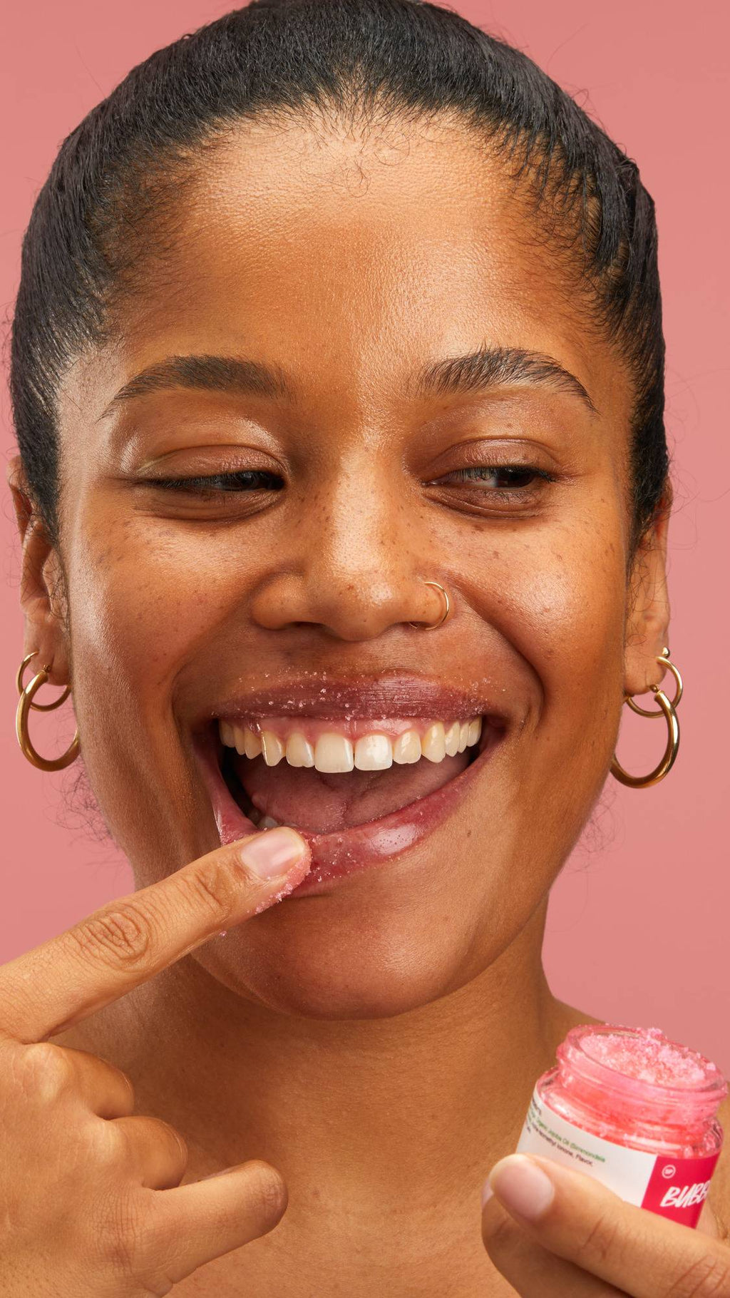 A model smiles while applying the Lush "Bubblegum" lip scrub to their bottom lip with a finger. They hold a small glass jar, which features a pink label with white text, in their other hand against a plain pink background.