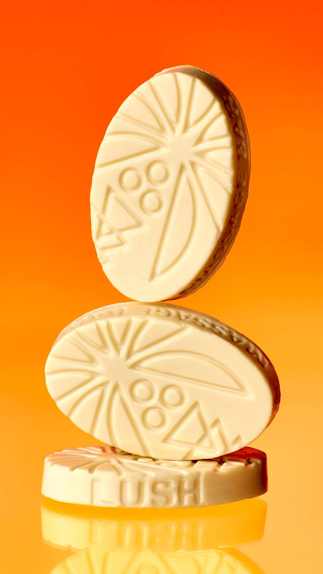 Three cream-coloured, patterned Lush "Coconut Drench" massage bars are stacked creatively on a glossy, reflective surface. The top bar is balanced vertically. The bottom bar displays the embossed word 'LUSH' against a vibrant orange background.