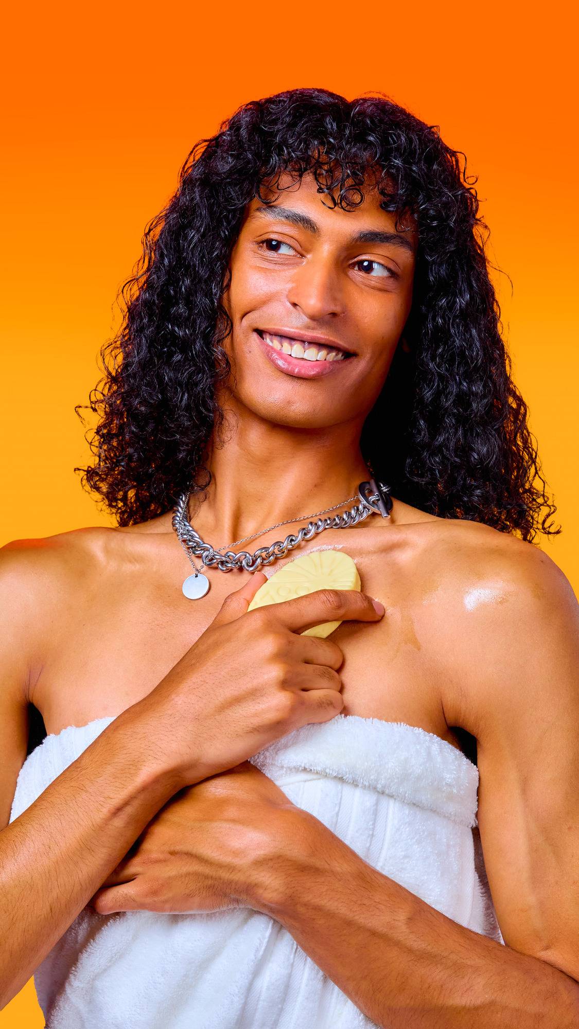 A smiling person with long, curly dark hair and a silver chain necklace holds a cream-coloured Lush massage bar against their collarbone. They are wrapped in a white towel, set against a bright orange background.