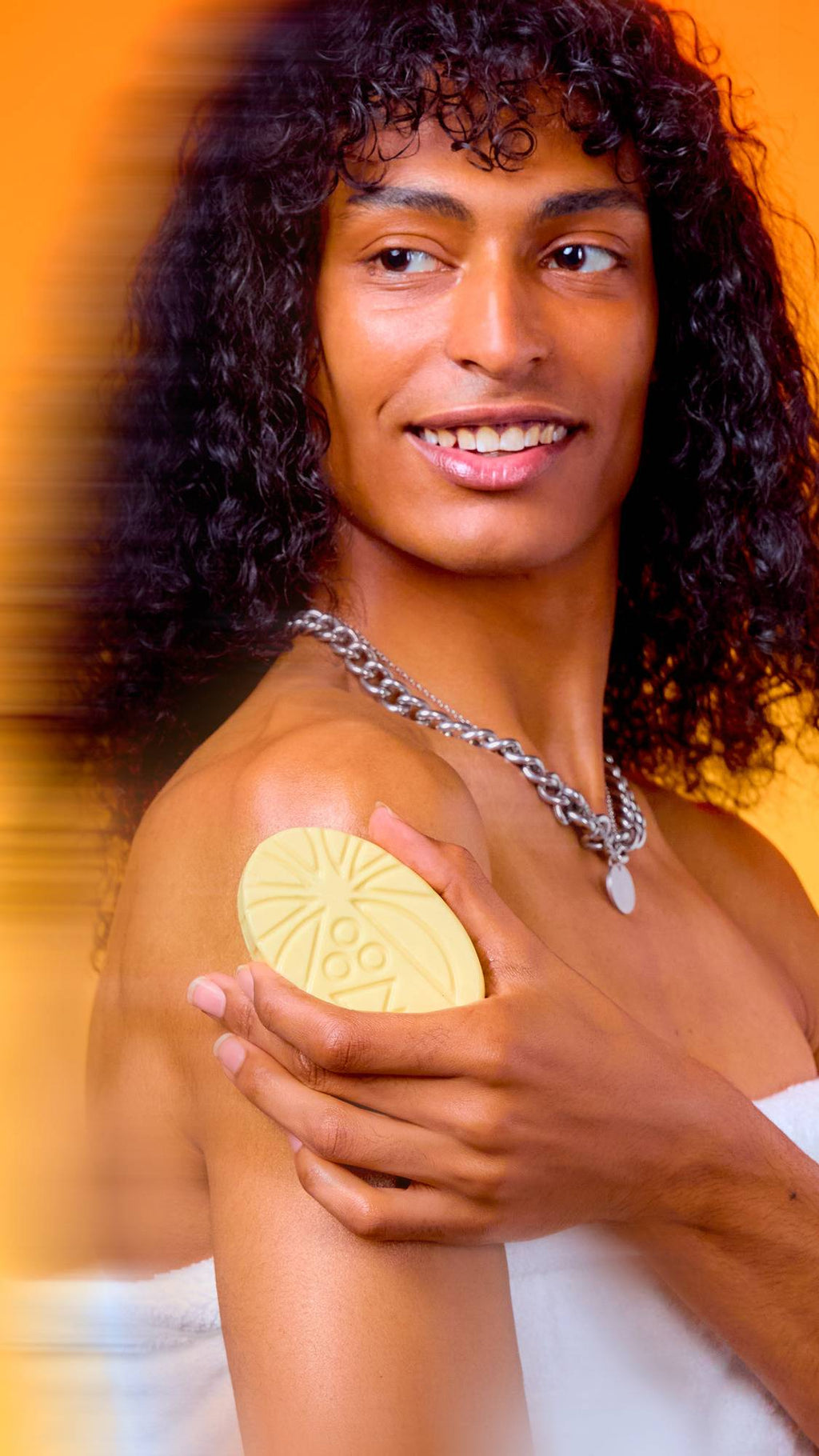 A smiling person with long, curly dark hair glides an oval, patterned cream Lush massage bar over their shoulder. They wear a silver chain necklace and a white towel, set against a warm, vibrant orange background.